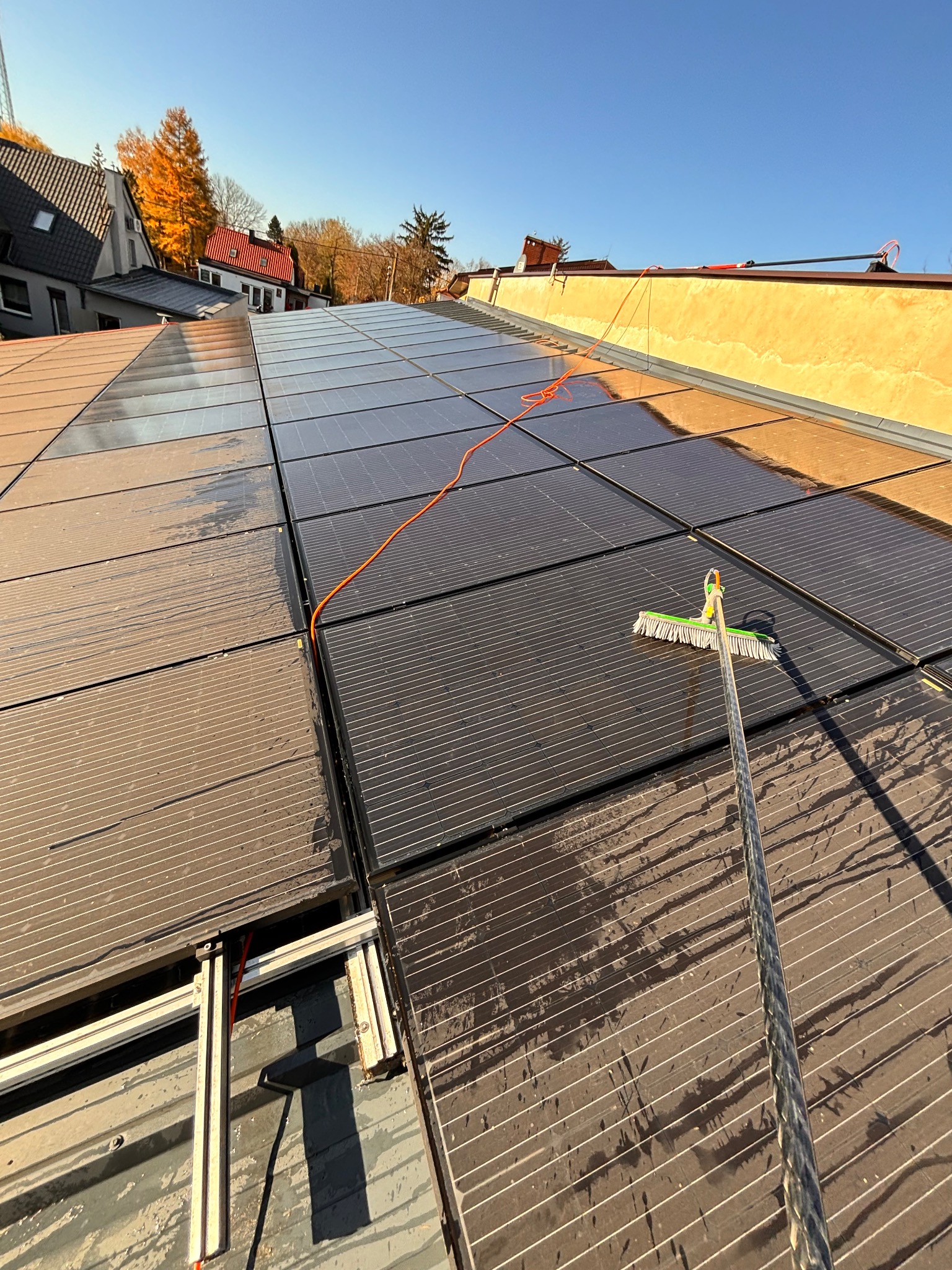 Czyszczenie paneli słonecznych na dachu domu za pomocą szczotki na długim kiju. Część paneli umyta, część zabrudzona. Widok z dołu na niebieskie niebo.