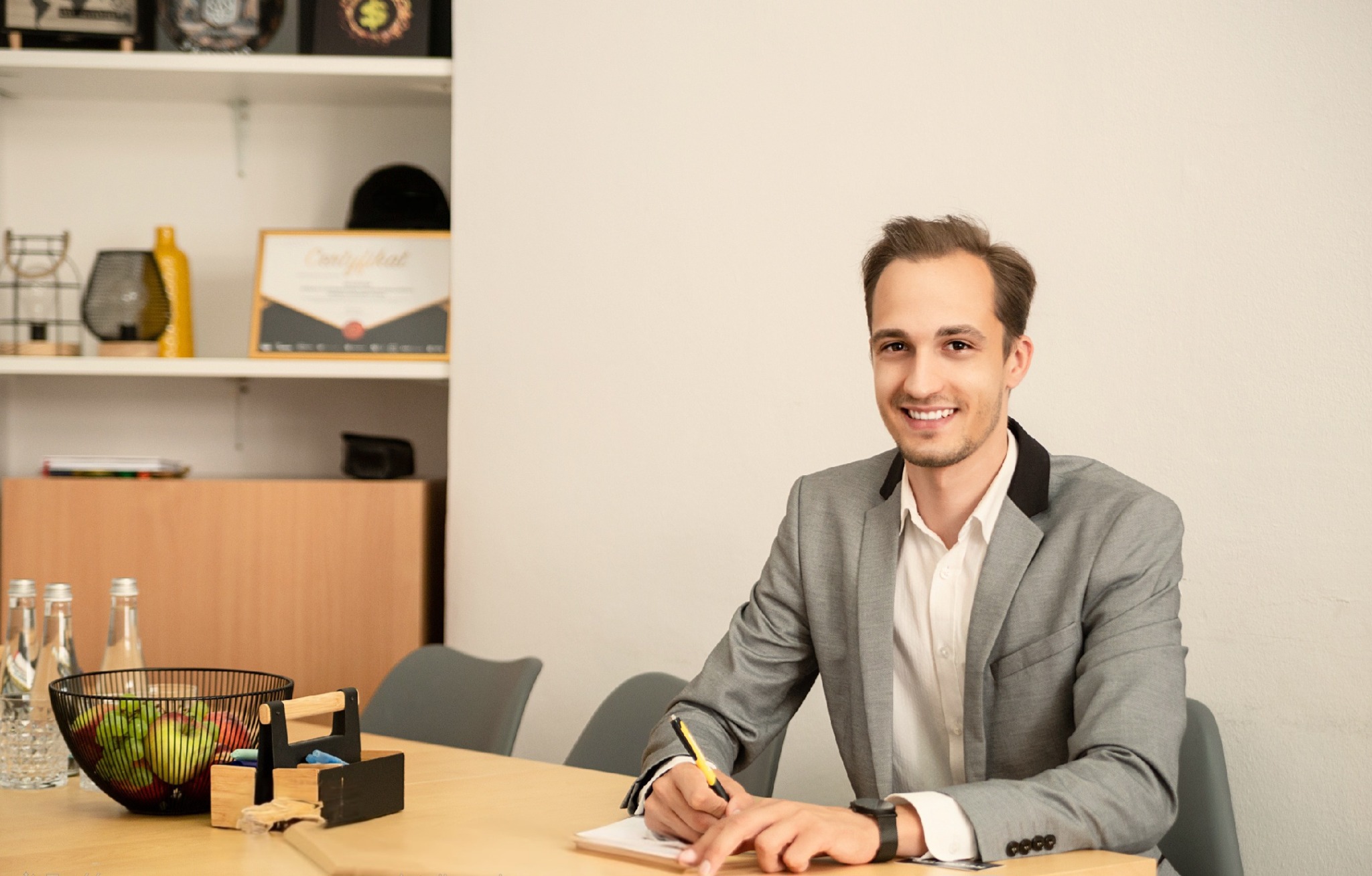 Uśmiechnięty mężczyzna w szarej marynarce siedzi przy biurku, pisząc coś na kartce żółtym ołówkiem. Na półce w tle widoczny certyfikat w złotej ramce.