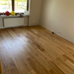 VipBud - Pokój z nową, jasną, dębową podłogą ułożoną w jodełkę, widoczny grzejnik pod oknem z dekoracyjnymi kulami i odsłonięte ściany przygotowane do malowania.