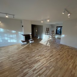 VipBud - Przestronne wnętrze po odświeżeniu: parkiet ułożony w jodełkę, białe ściany z widocznymi śladami światła słonecznego, dwa drewniane drabiny malarskie w głębi pomieszczenia i nowoczesny kominek.