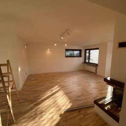 VipBud - Jasne, przestronne wnętrze z drewnianą podłogą i białymi ścianami, oświetlone światłem słonecznym wpadającym przez okna, z widoczną drabiną malarską po lewej stronie i kominkiem po prawej...