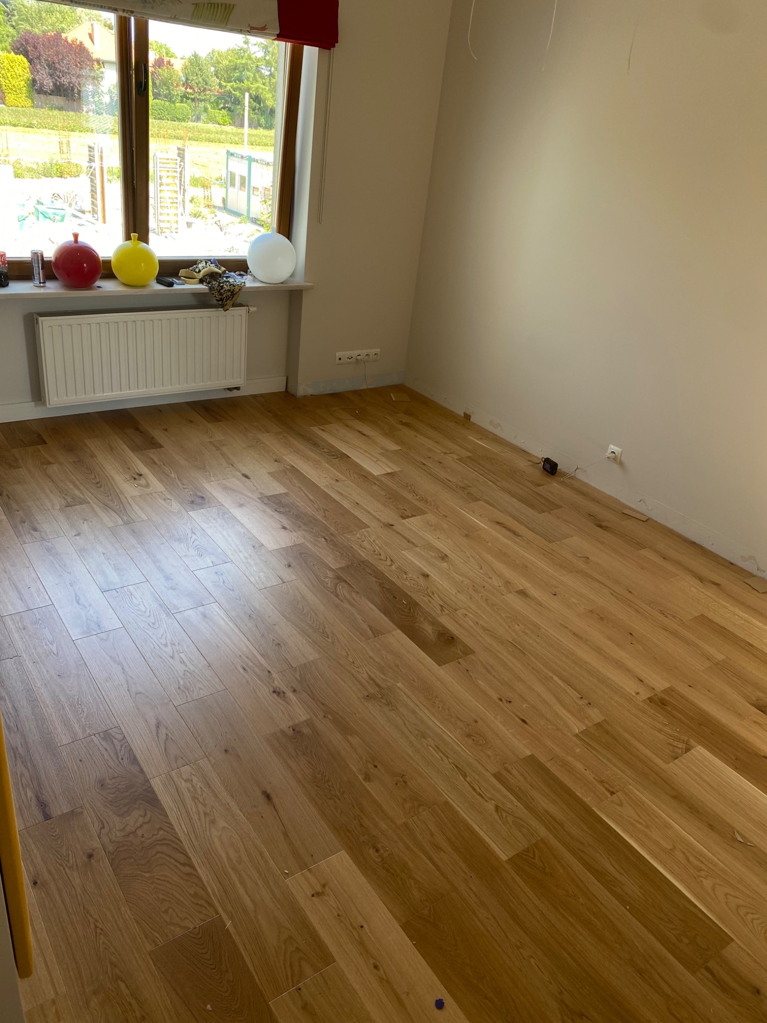 Pokój z nową, jasną, dębową podłogą ułożoną w jodełkę, widoczny grzejnik pod oknem z dekoracyjnymi kulami i odsłonięte ściany przygotowane do malowania.