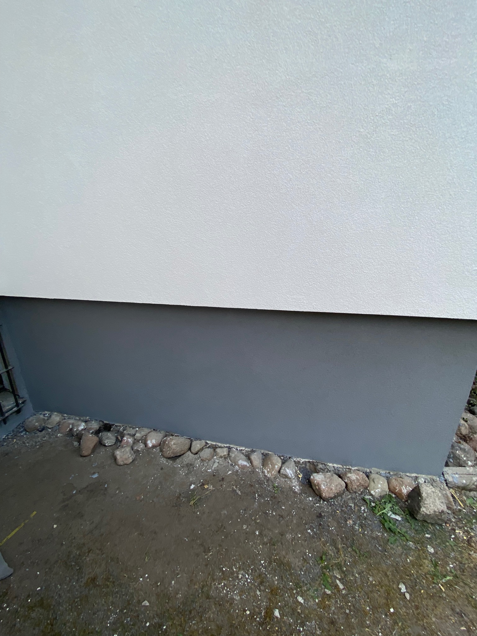 Fragment ściany zewnętrznej budynku z pomalowaną szarą farbą dolną partią, odgrodzoną od ziemi rzędem kamieni.