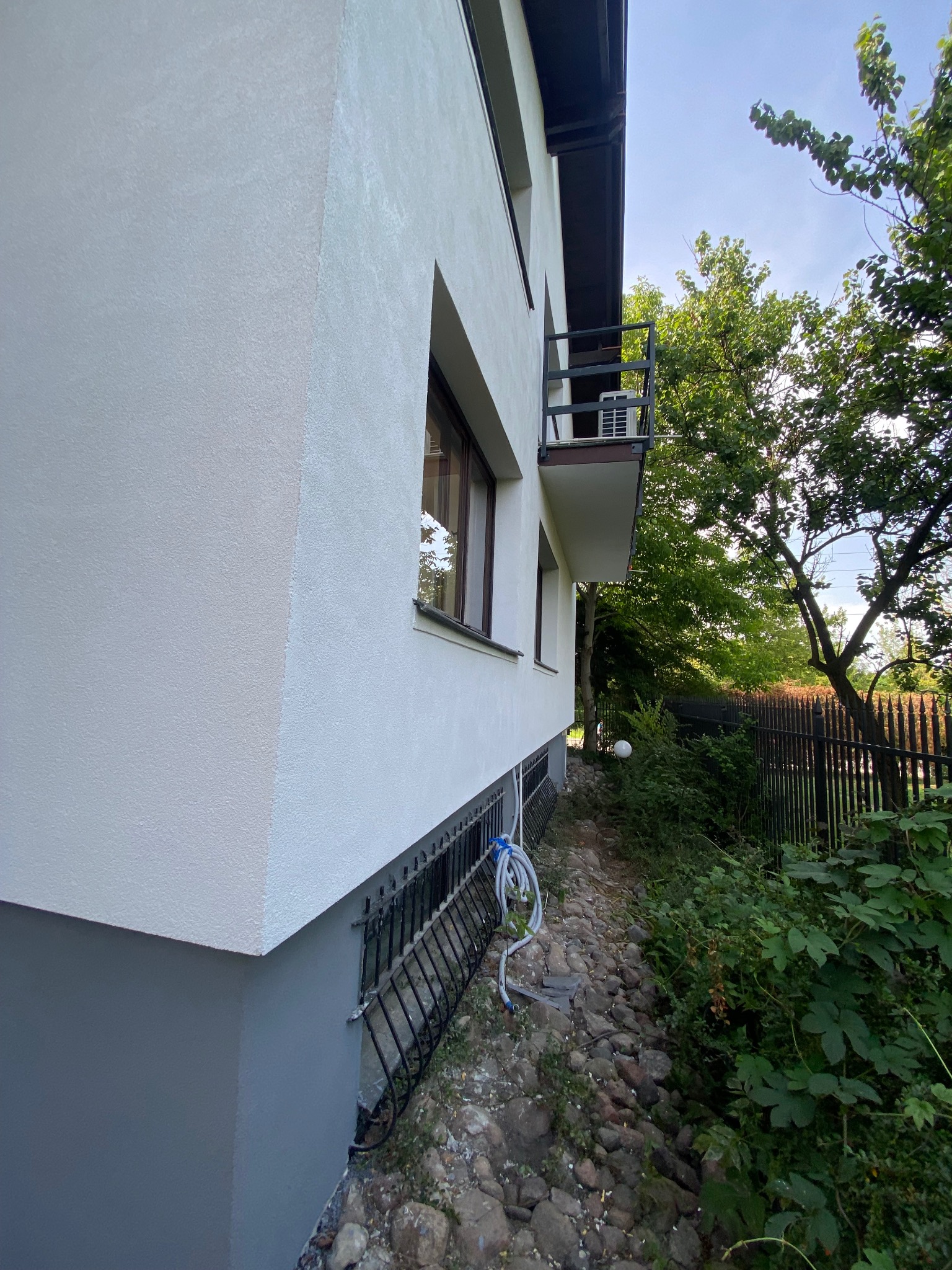 Świeżo pomalowana elewacja domu z widoczną fakturą tynku, fragment balkonu z klimatyzatorem oraz roślinność przy ogrodzeniu.