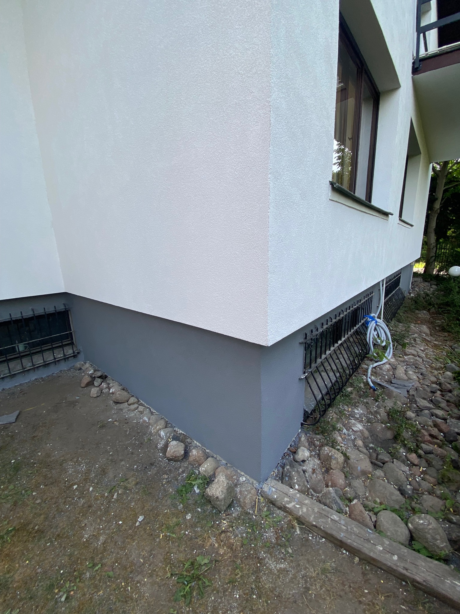 Odnowiona elewacja domu z pomalowanym na szaro cokołem, widoczne okratowane okna piwniczne, biała ściana z delikatną fakturą i fragment zwiniętego węża ogrodowego.