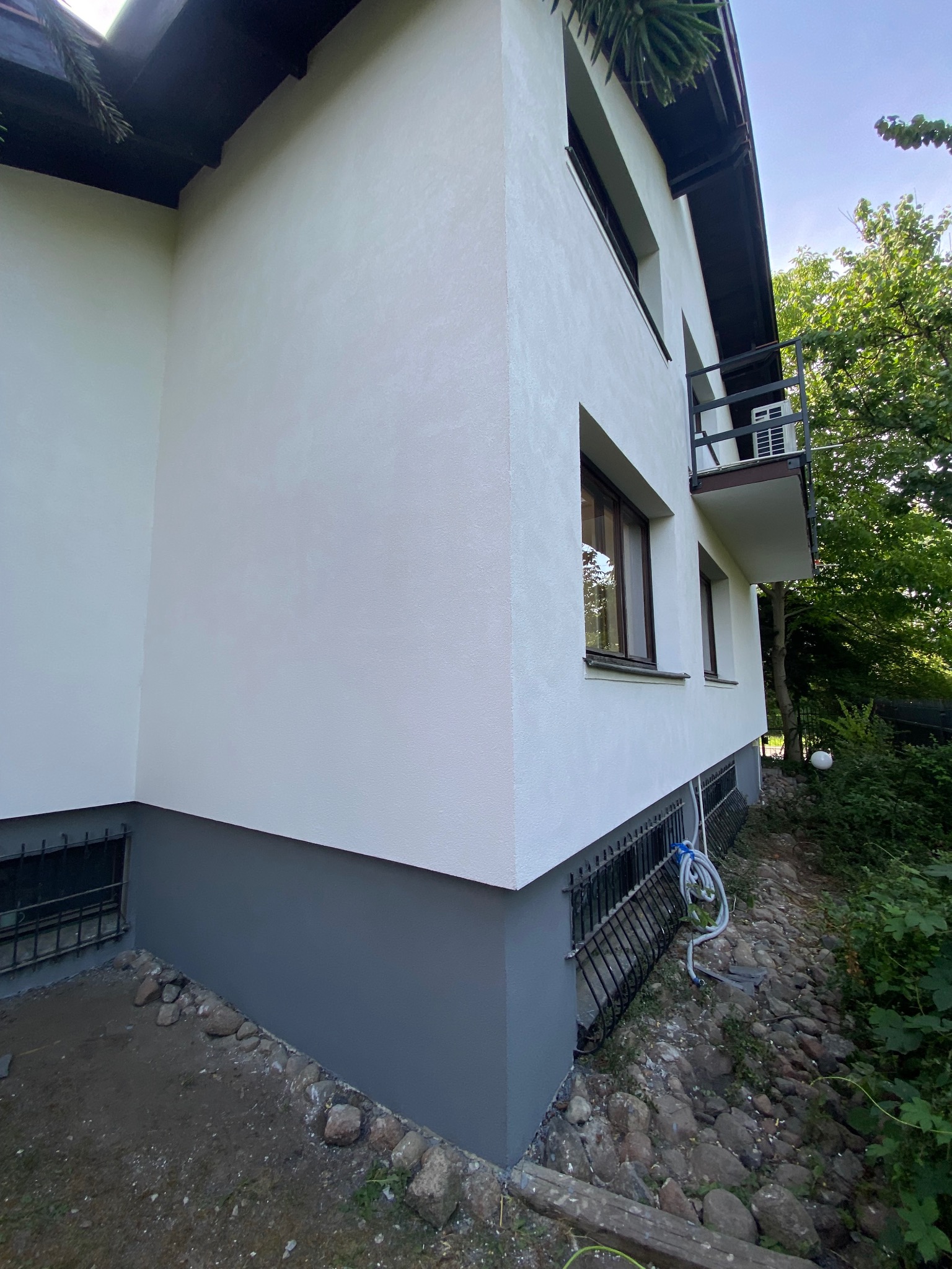 Fragment elewacji domu po malowaniu, z widocznym cokołem w kolorze szarym i świeżo pomalowaną białą fasadą, balkon z klimatyzatorem, okna z brązowymi ramami, teren wokół domu z kamieniami...
