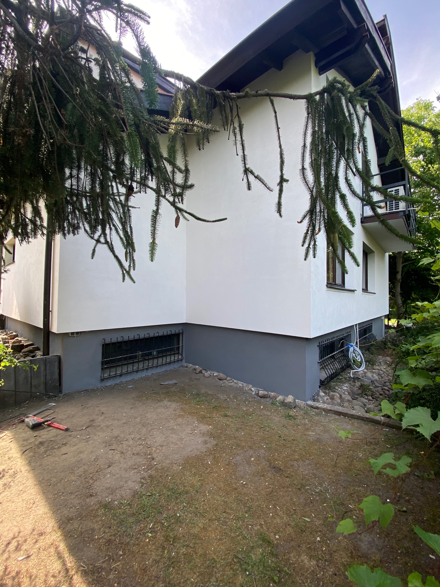 Odnowiona elewacja domu w Warszawie z pomalowaną na szaro podmurówką, widoczne narzędzia na ziemi i gałąź drzewa w kadrze.