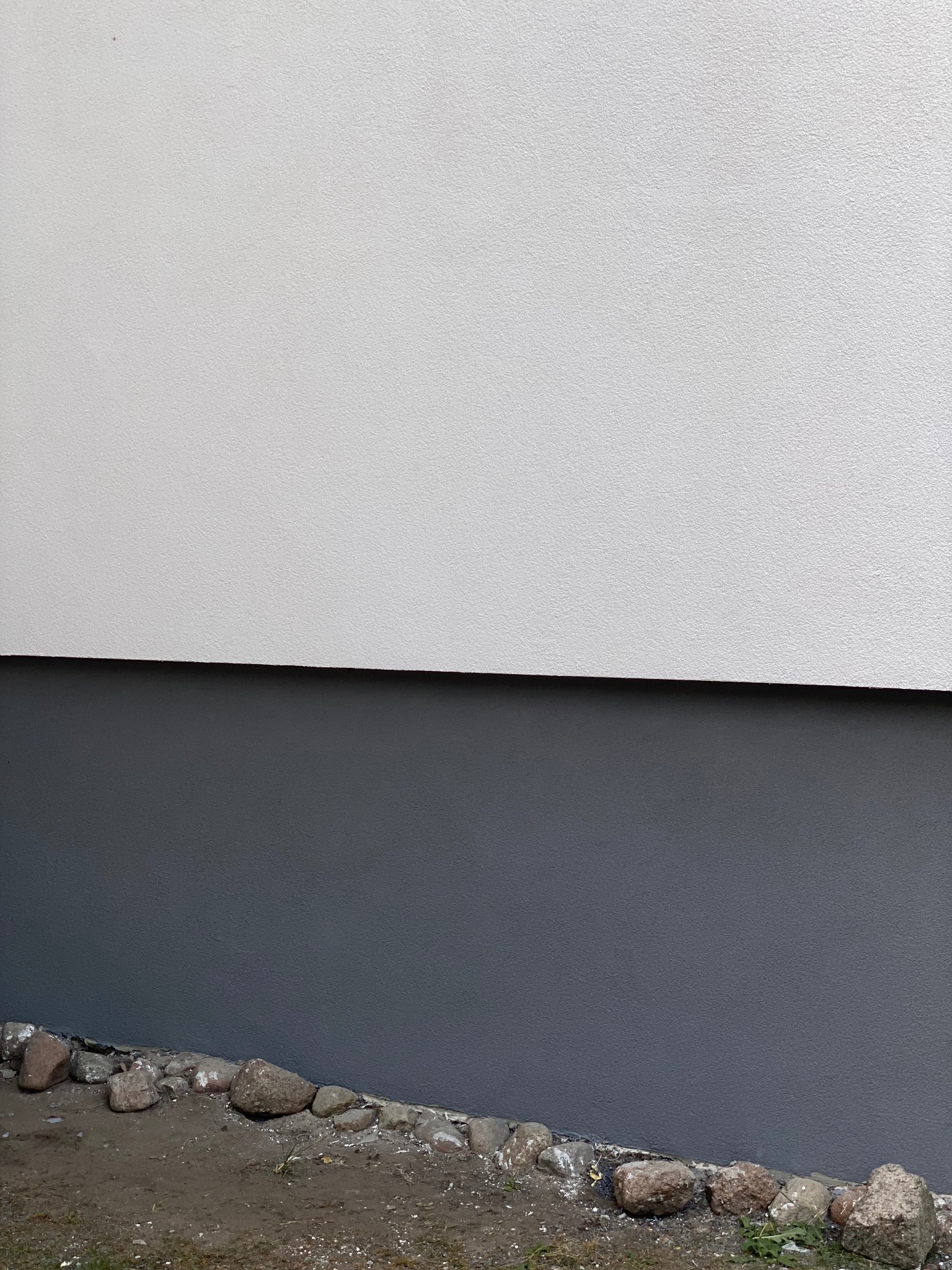 Dwukolorowa elewacja budynku z szarą dolną częścią i białym tynkiem powyżej, oddzielona cienką czarną linią, z kamieniami u podstawy.