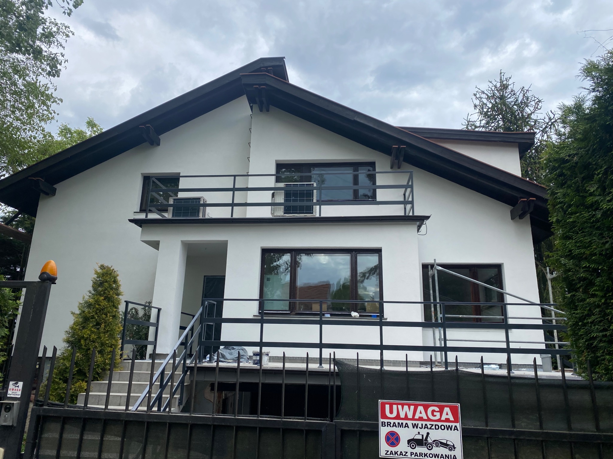Biały dom jednorodzinny z ciemnobrązowym dachem i balkonem, widoczny zza czarnego metalowego ogrodzenia z tabliczką 'Uwaga Brama Wjazdowa', częściowo zasłonięty rusztowaniem przy oknie.
