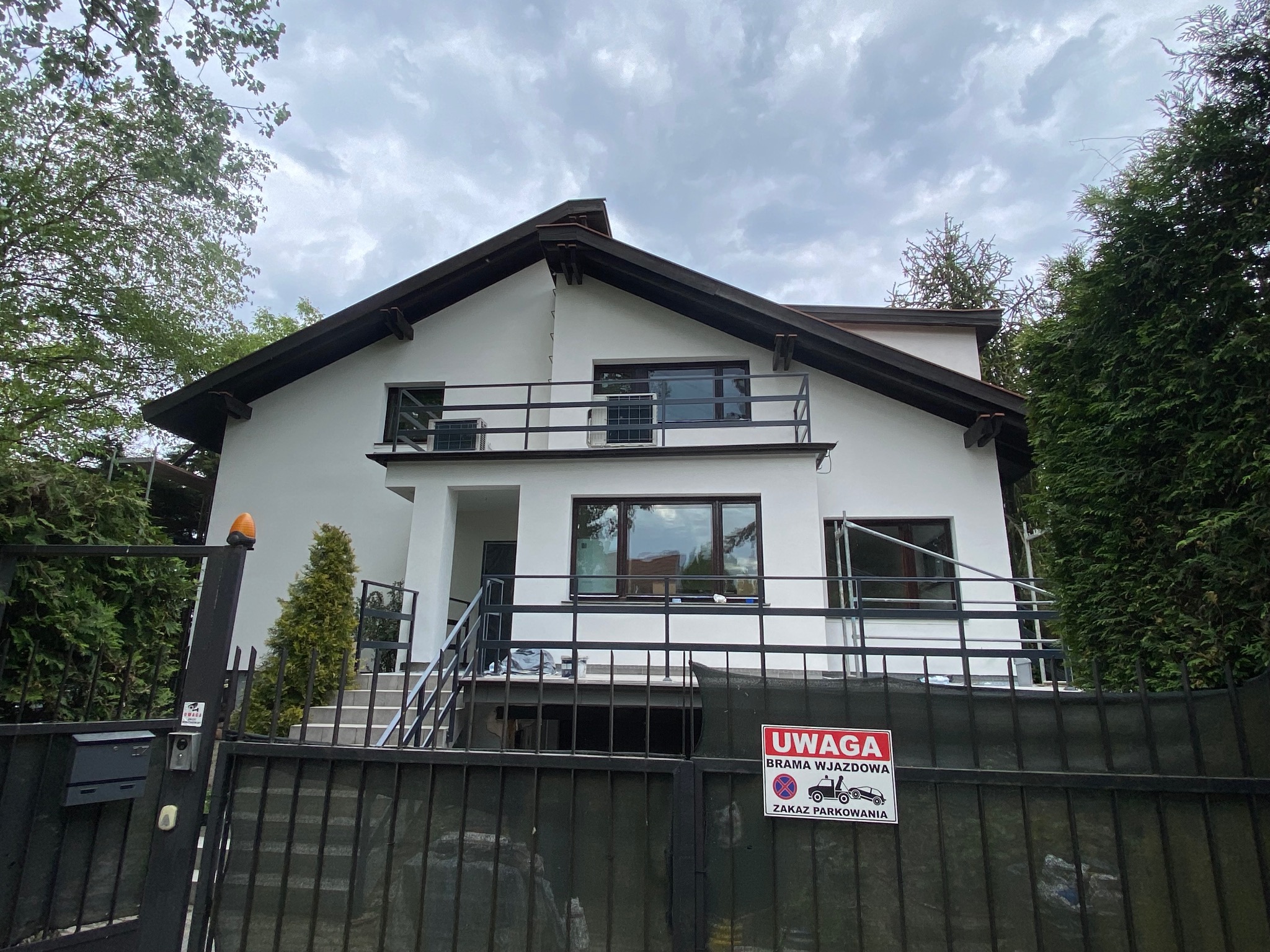 Dwupiętrowy dom z balkonem, świeżo pomalowany na biało, widoczny zza czarnej bramy z tabliczką 'Uwaga brama wjazdowa - zakaz parkowania'.