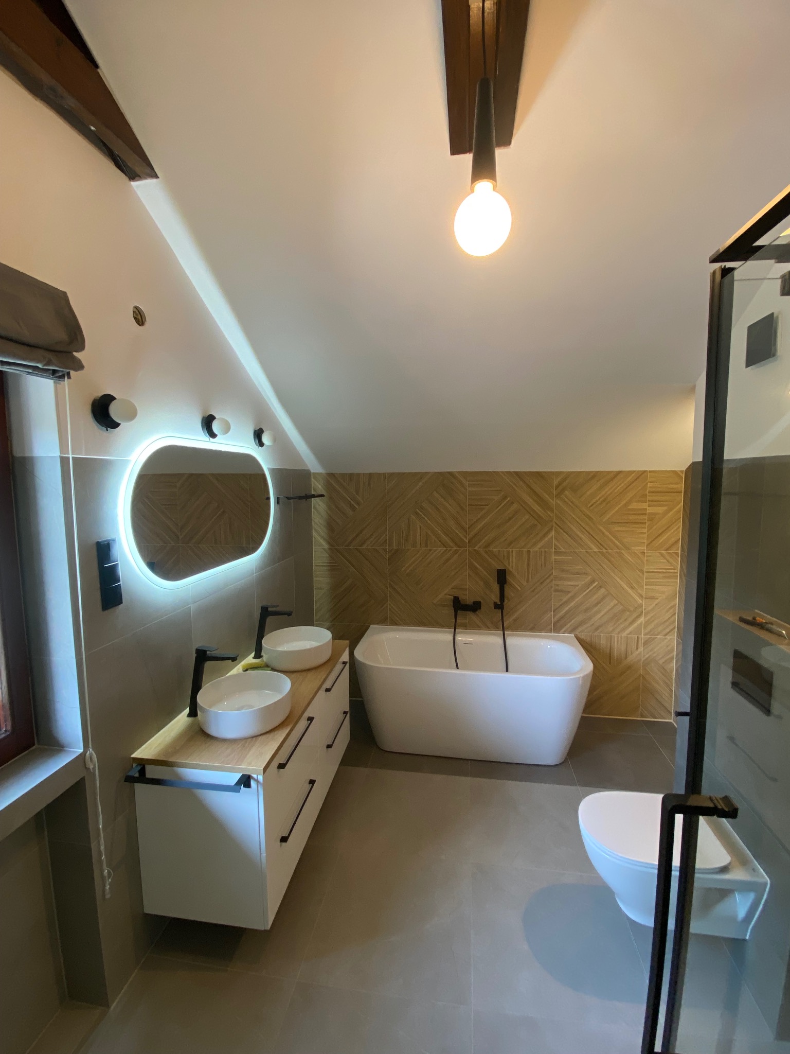 Nowoczesna łazienka na poddaszu z wolnostojącą wanną, podświetlanym lustrem, drewnopodobnymi płytkami na ścianie i czarną armaturą.