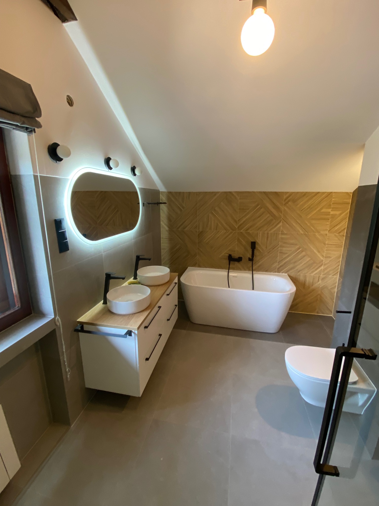 Nowoczesna łazienka na poddaszu z dwoma umywalkami nablatowymi na drewnianym blacie, podświetlanym lustrem, wolnostojącą wanną i toaletą, wykończona płytkami imitującymi drewno na ścianie.