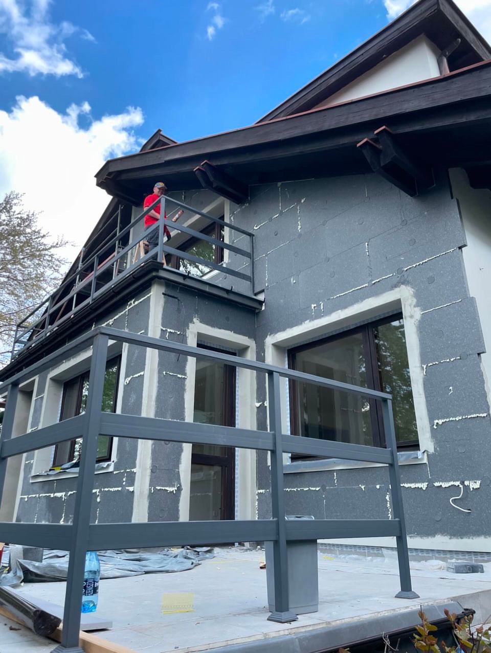 Ocieplanie elewacji domu jednorodzinnego: szare płyty styropianowe z widocznymi szczelinami wypełnionymi pianką montażową, pracownik w czerwonej koszulce na balkonie