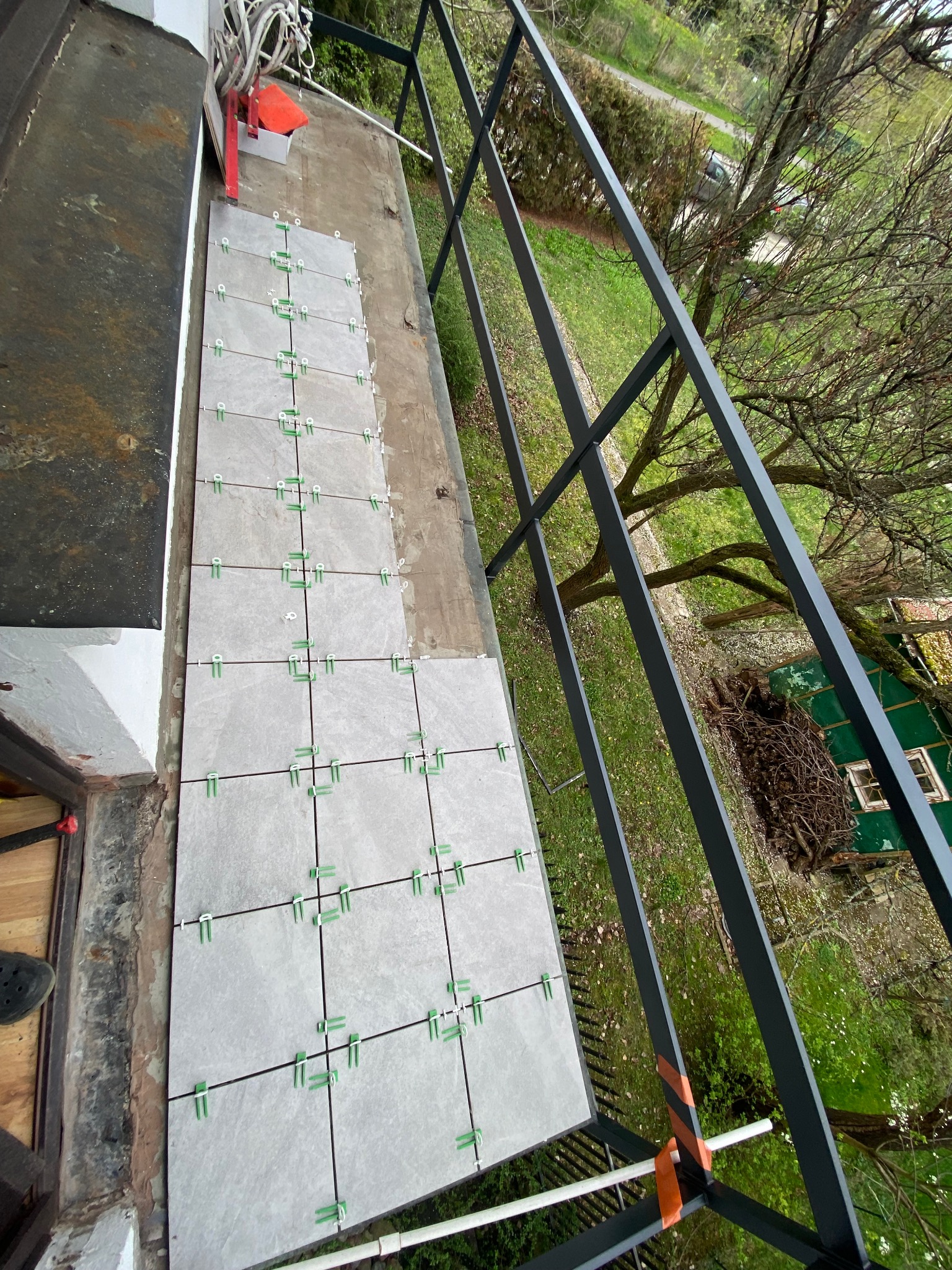 Układanie szarych płytek na balkonie z użyciem zielonych krzyżyków dystansowych, widok z góry przez czarną balustradę na drzewa i zielony teren.