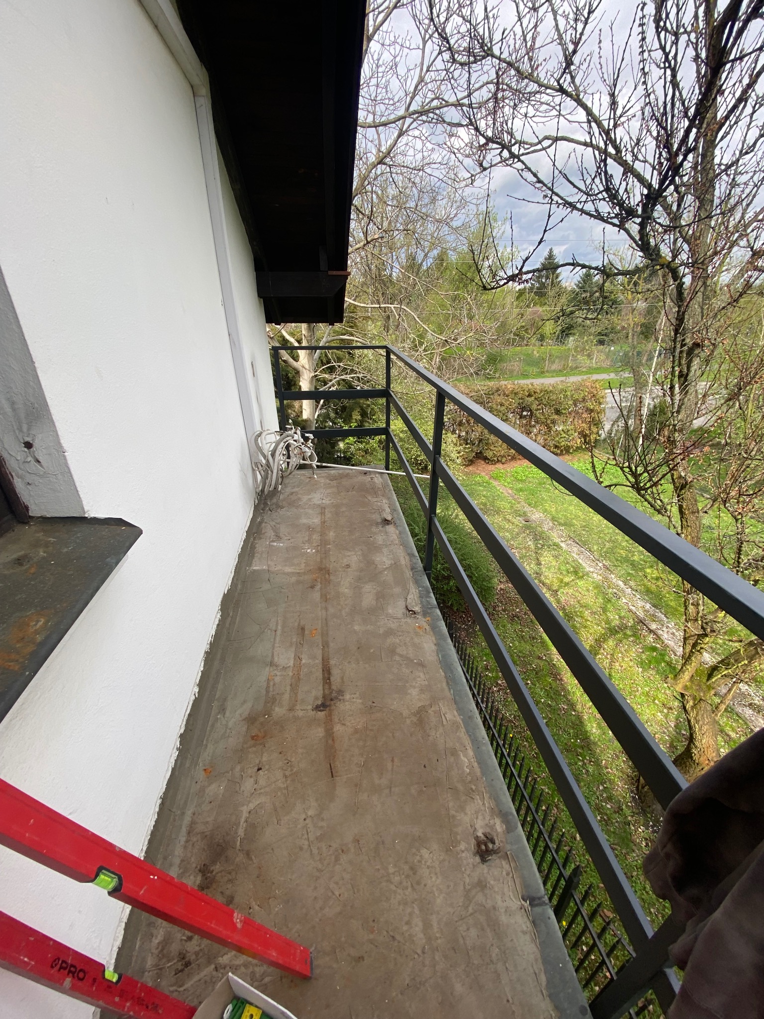 Betonowy balkon przed renowacją z widocznym poziomem, niepomalowaną ścianą i balustradą z metalowych prętów, widok na drzewa i zieleń.