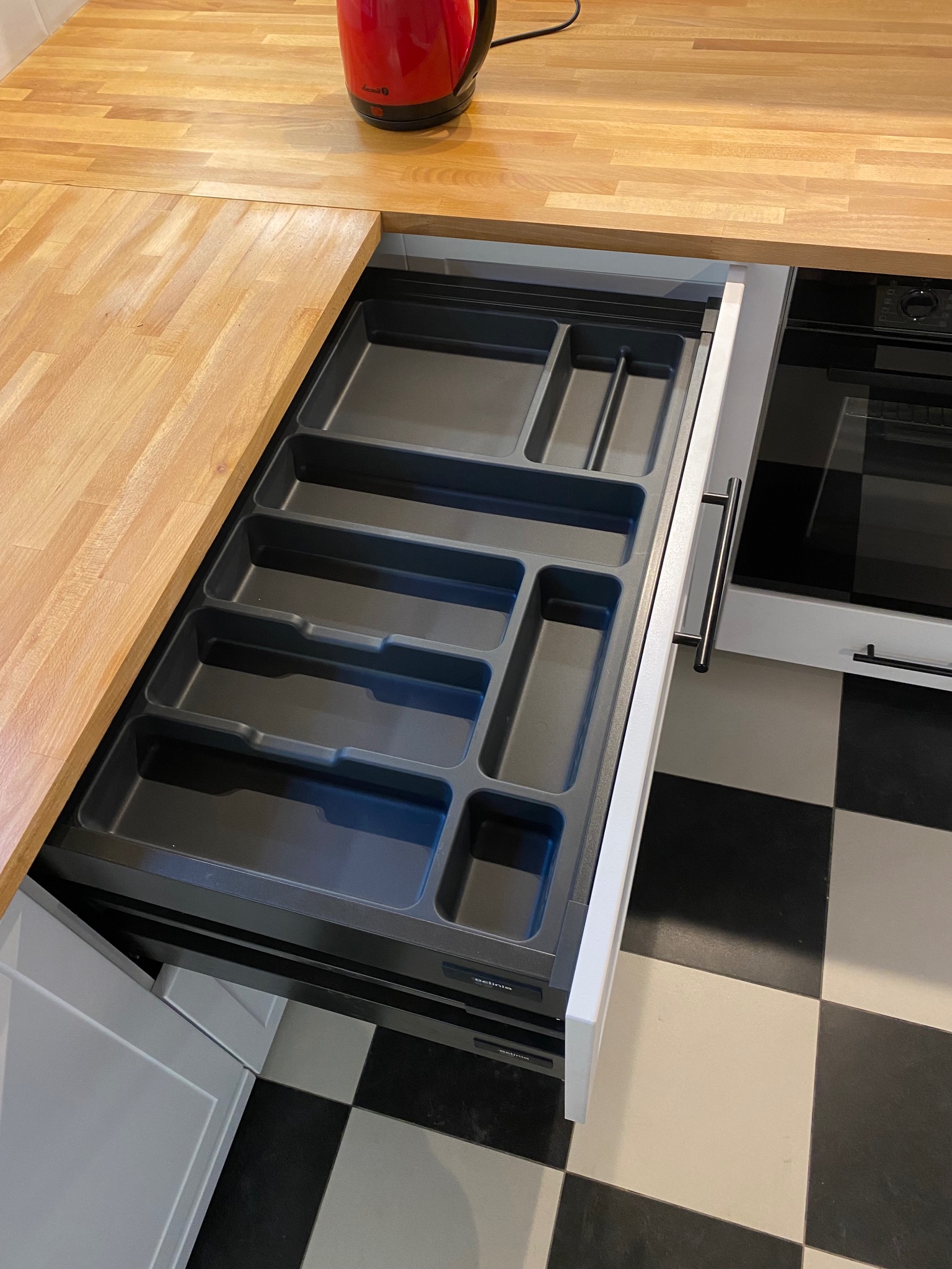 Otwarta szuflada kuchenna z szarym wkładem na sztućce i akcesoria, widoczny fragment drewnianego blatu i piekarnika, podłoga w czarno-białą szachownicę.