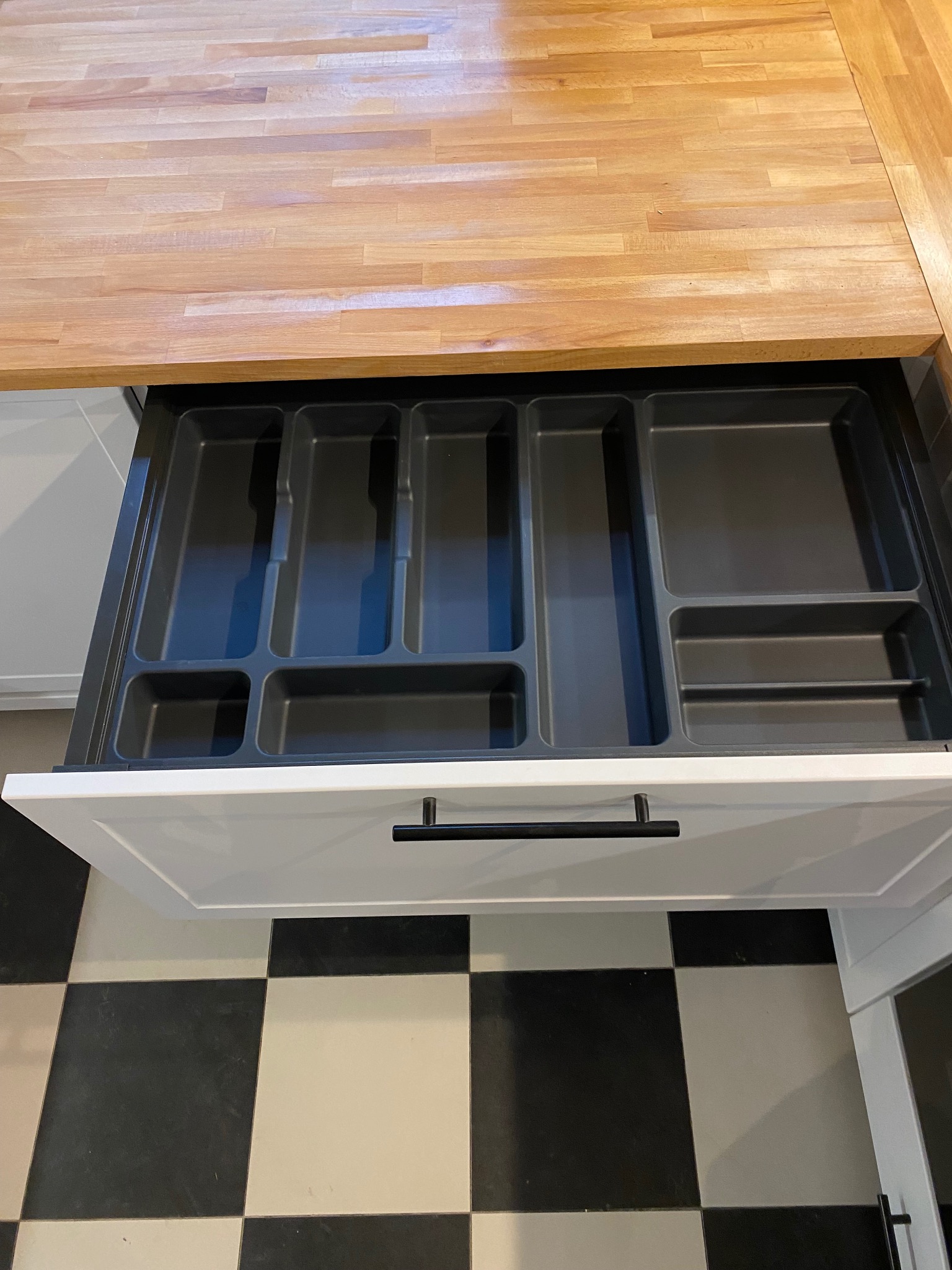 Otwarta szuflada kuchenna z organizerem na sztućce i akcesoria, widoczna drewniana powierzchnia blatu i czarno-biała podłoga w szachownicę.