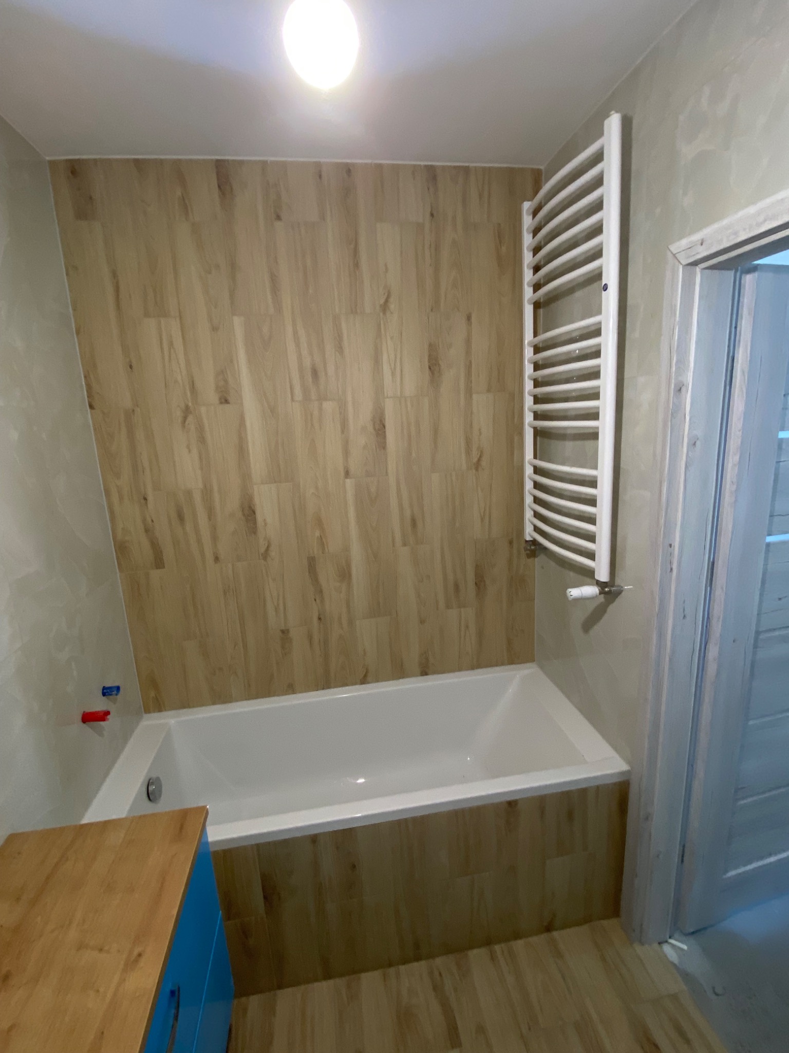 Nowoczesna łazienka z wanną obudowaną panelami imitującymi drewno, grzejnikiem drabinkowym i szafką z niebieskimi frontami.