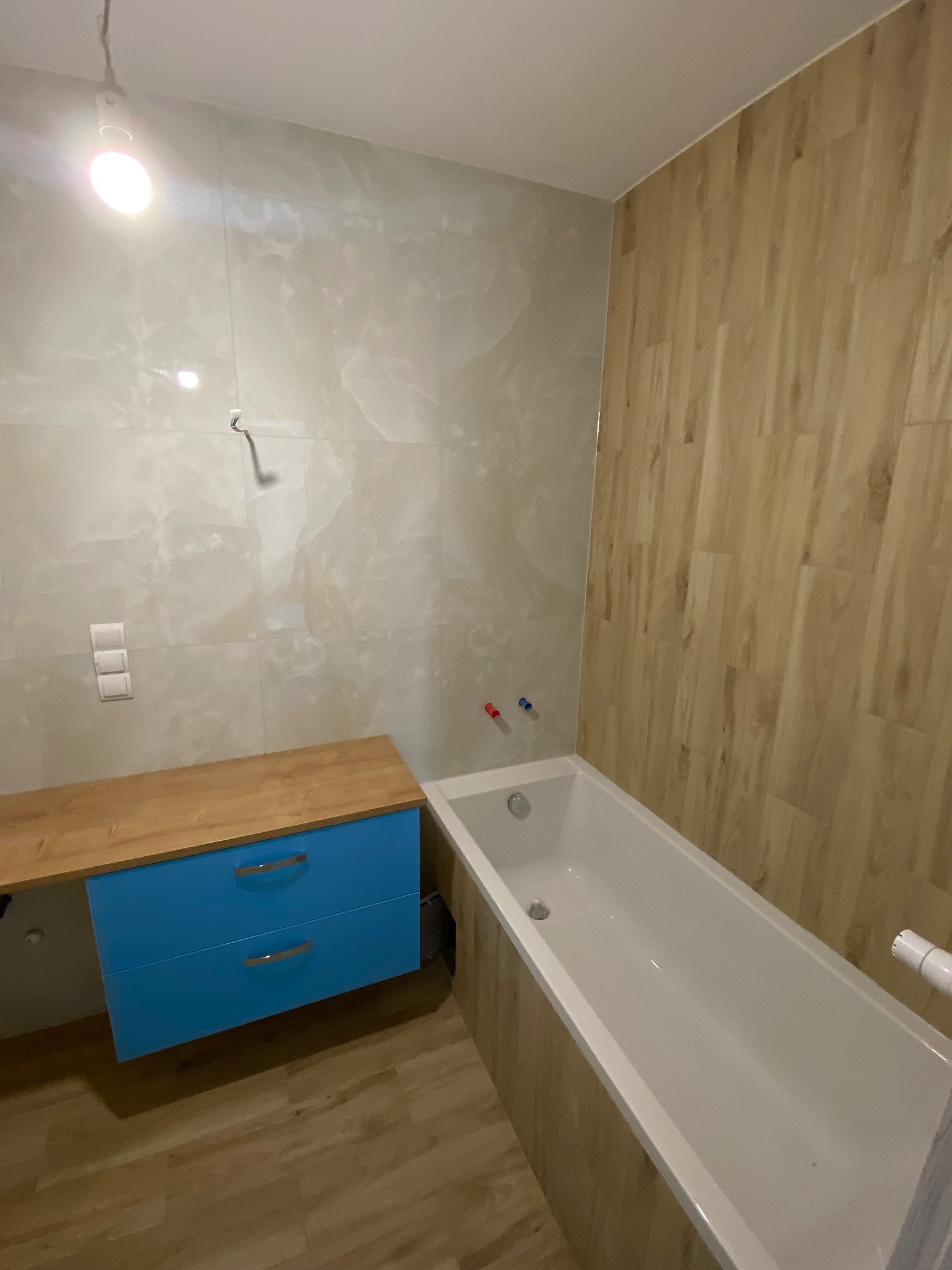 Nowoczesna łazienka z drewnopodobnymi płytkami ściennymi, białą wanną, błękitną szafką pod umywalką z drewnianym blatem i przełącznikami na ścianie w tle.