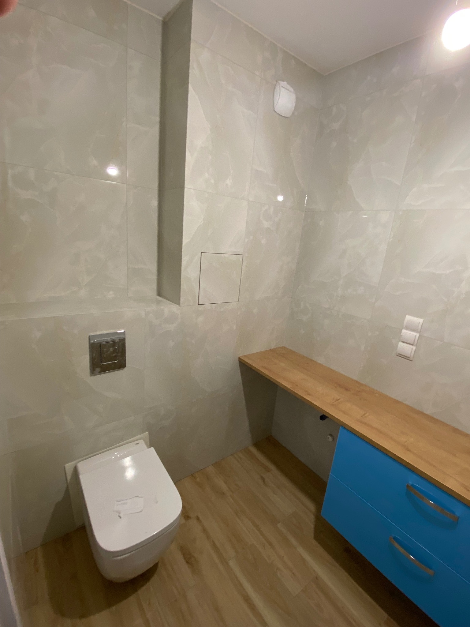 Wykończona łazienka z podwieszaną toaletą, drewnopodobnymi płytkami podłogowymi, szarymi marmurkowymi płytkami ściennymi i błękitną szafką pod umywalkę z drewnianym blatem.