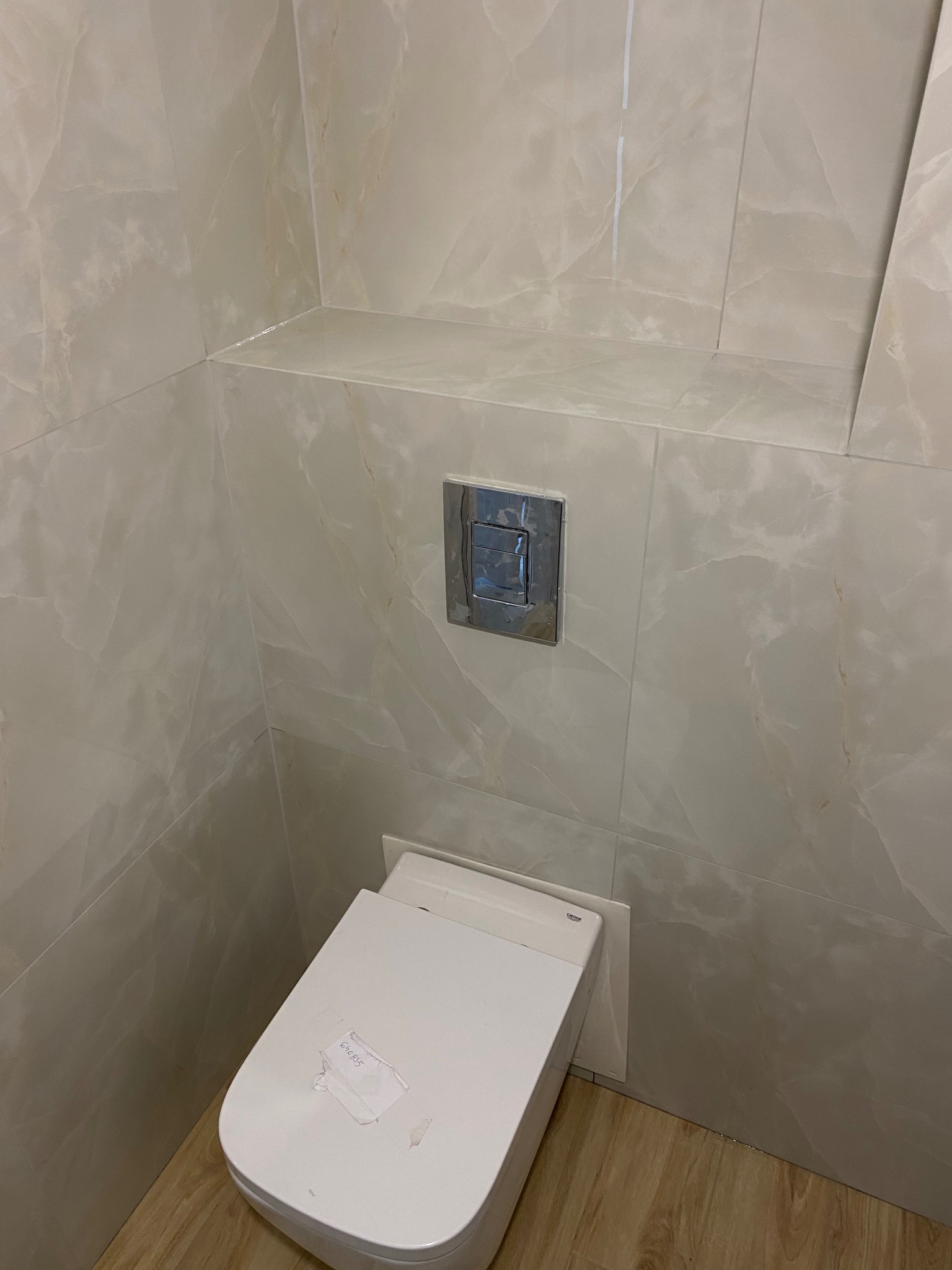 Wisząca toaleta z przyciskiem spłukującym w łazience wykończonej płytkami imitującymi marmur, widoczne niedokładności montażu w postaci naklejki na desce sedesowej i niedoczyszczonych fug.