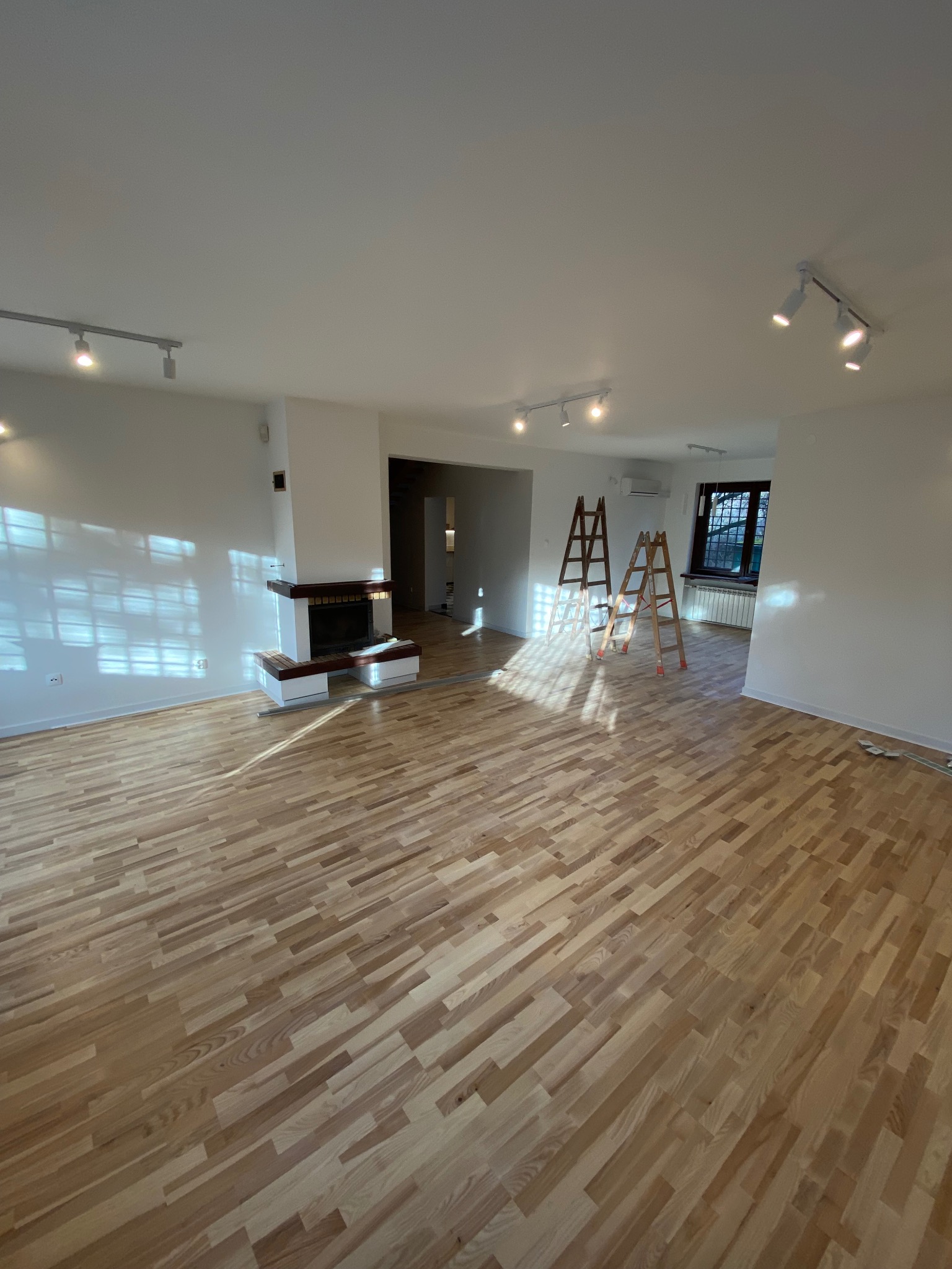 Przestronne wnętrze po odświeżeniu: parkiet ułożony w jodełkę, białe ściany z widocznymi śladami światła słonecznego, dwa drewniane drabiny malarskie w głębi pomieszczenia i nowoczesny kominek.