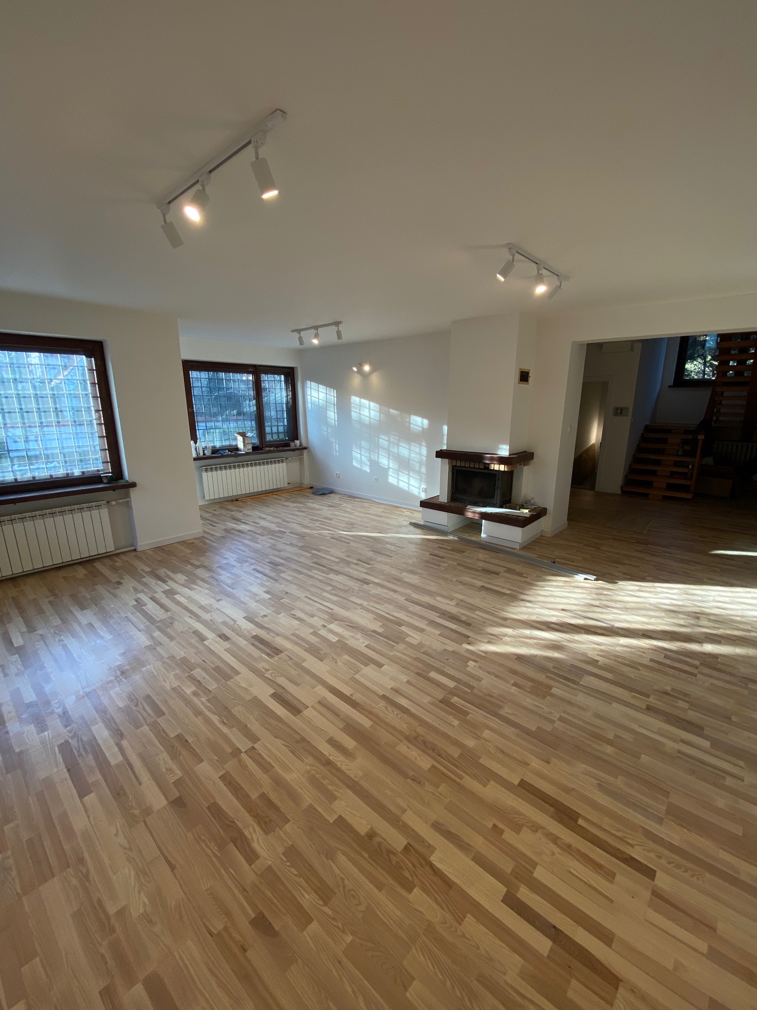 Jasny, przestronny salon z parkietem w jodełkę, białymi ścianami, nowoczesnym oświetleniem punktowym, oknami z kratami i kominkiem. Na podłodze leżą listwy przypodłogowe. W głębi widoczne schody.