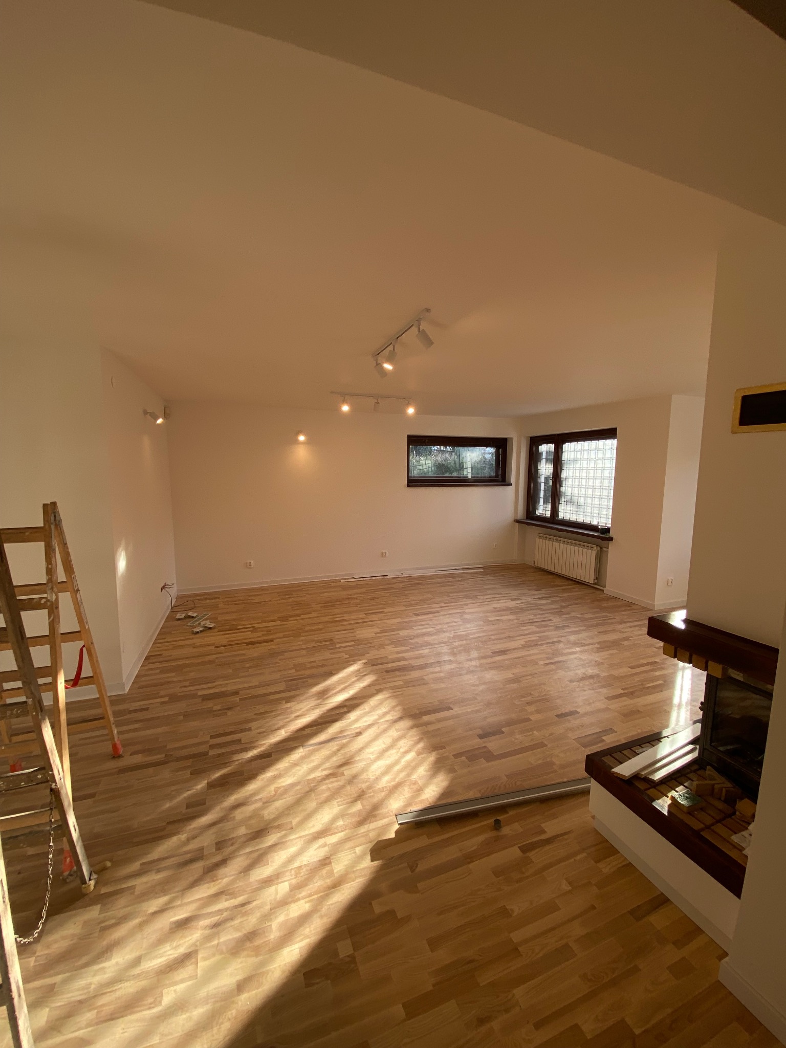 Jasne, przestronne wnętrze z drewnianą podłogą i białymi ścianami, oświetlone światłem słonecznym wpadającym przez okna, z widoczną drabiną malarską po lewej stronie i kominkiem po prawej...