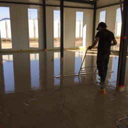 SIGMA MARIUSZ DOBROWOLSKI - Wylewka anhydrytowa w jasnym pomieszczeniu z oknami. Pracownik rozprowadza masę poziomującą. Widoczne elementy ogrzewania podłogowego i pionowe znaczniki poziomu.