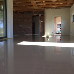 SIGMA MARIUSZ DOBROWOLSKI - Świeżo wylana, gładka posadzka anhydrytowa w surowym wnętrzu z widocznymi elementami konstrukcyjnymi i dużymi oknami, odbijającymi światło.