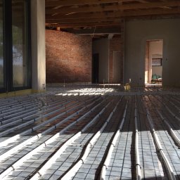SIGMA MARIUSZ DOBROWOLSKI - Instalacja ogrzewania podłogowego w surowym wnętrzu z ceglaną ścianą. Rury grzewcze ułożone na macie, przygotowane do wylewki. Widoczne okno i surowe wykończenie.