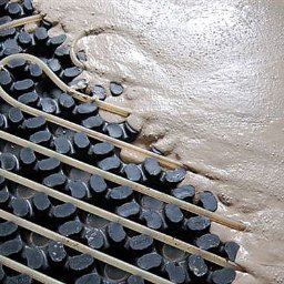 SIGMA MARIUSZ DOBROWOLSKI - Proces zalewania rur ogrzewania podłogowego wylewką. Widoczne rury ułożone na czarnej macie i świeża, jasna wylewka pokrywająca część powierzchni.