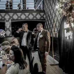 Dwaj mężczyźni w garniturach piją wino podczas uroczystości, w tle dekoracja z kwiatów i czarnej tkaniny z neonowym napisem, widoczny stół z zastawą i świecami.