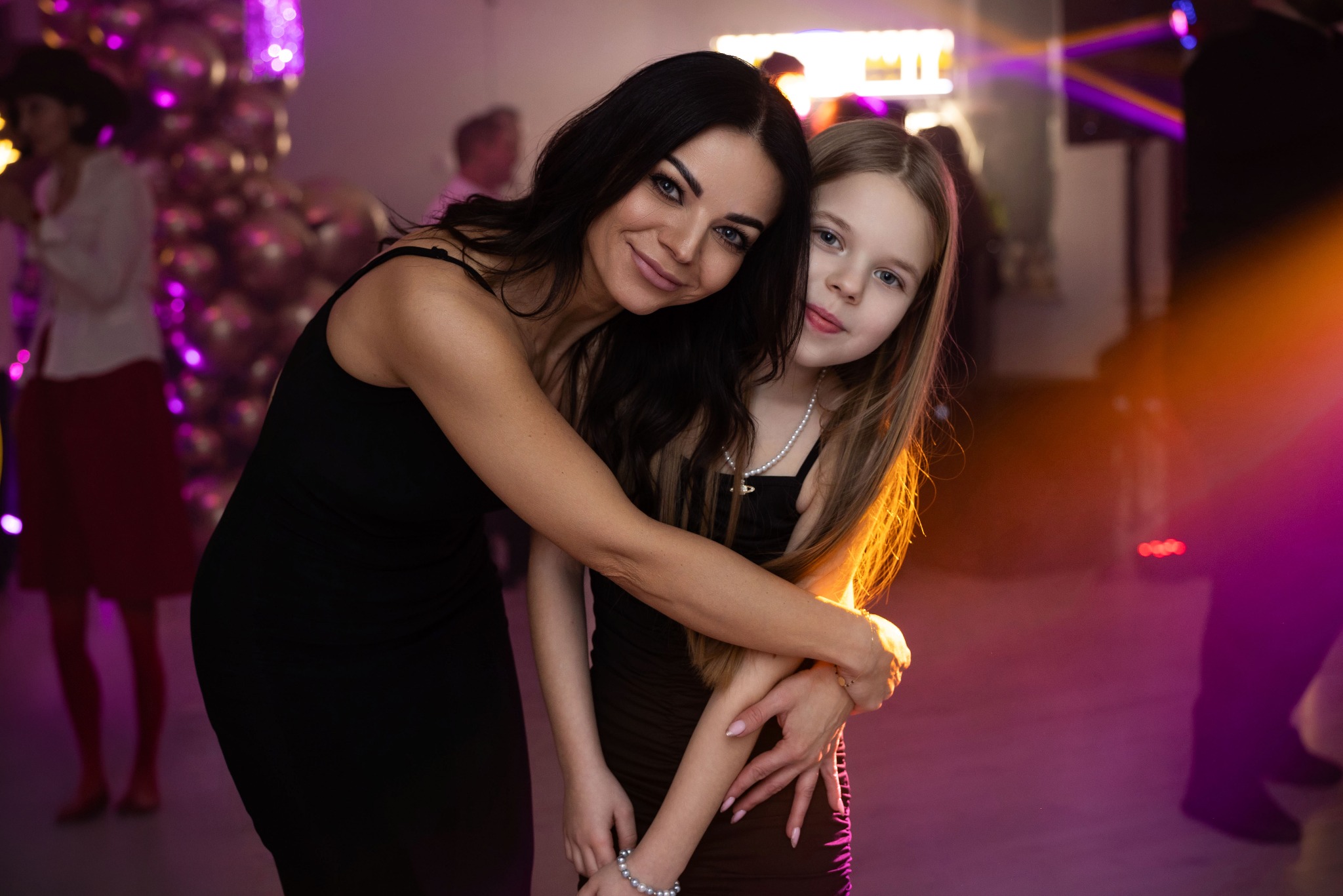 Kobieta obejmuje nastoletnią córkę na tle dekoracji z balonów i świateł podczas wydarzenia.