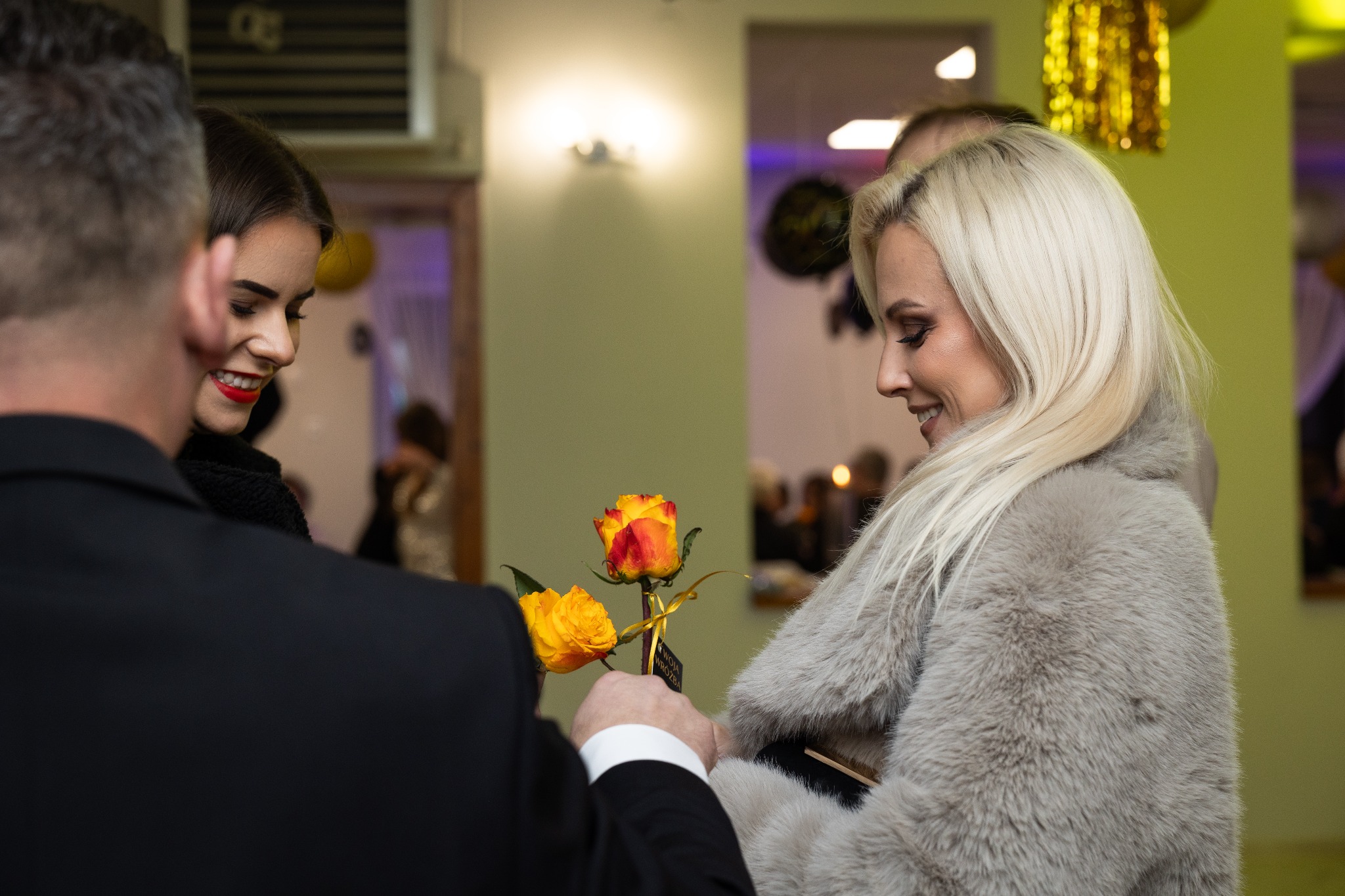 Mężczyzna w garniturze wręcza bukiet żółto-pomarańczowych róż uśmiechniętej blondynce w futrzanym płaszczu, w tle uśmiechnięta brunetka z czerwoną szminką i dekoracje imprezowe.