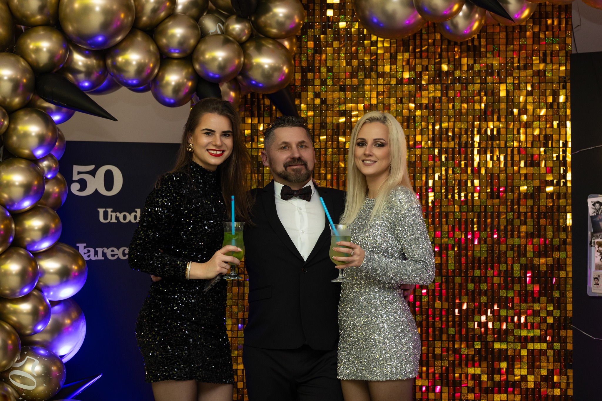 Trzy osoby pozują z drinkami na tle błyszczącej ściany i dekoracji balonowych z okazji 50. urodzin. Uśmiechnięta brunetka w czarnej sukience, mężczyzna w garniturze z muchą i blondynka w srebrnej...