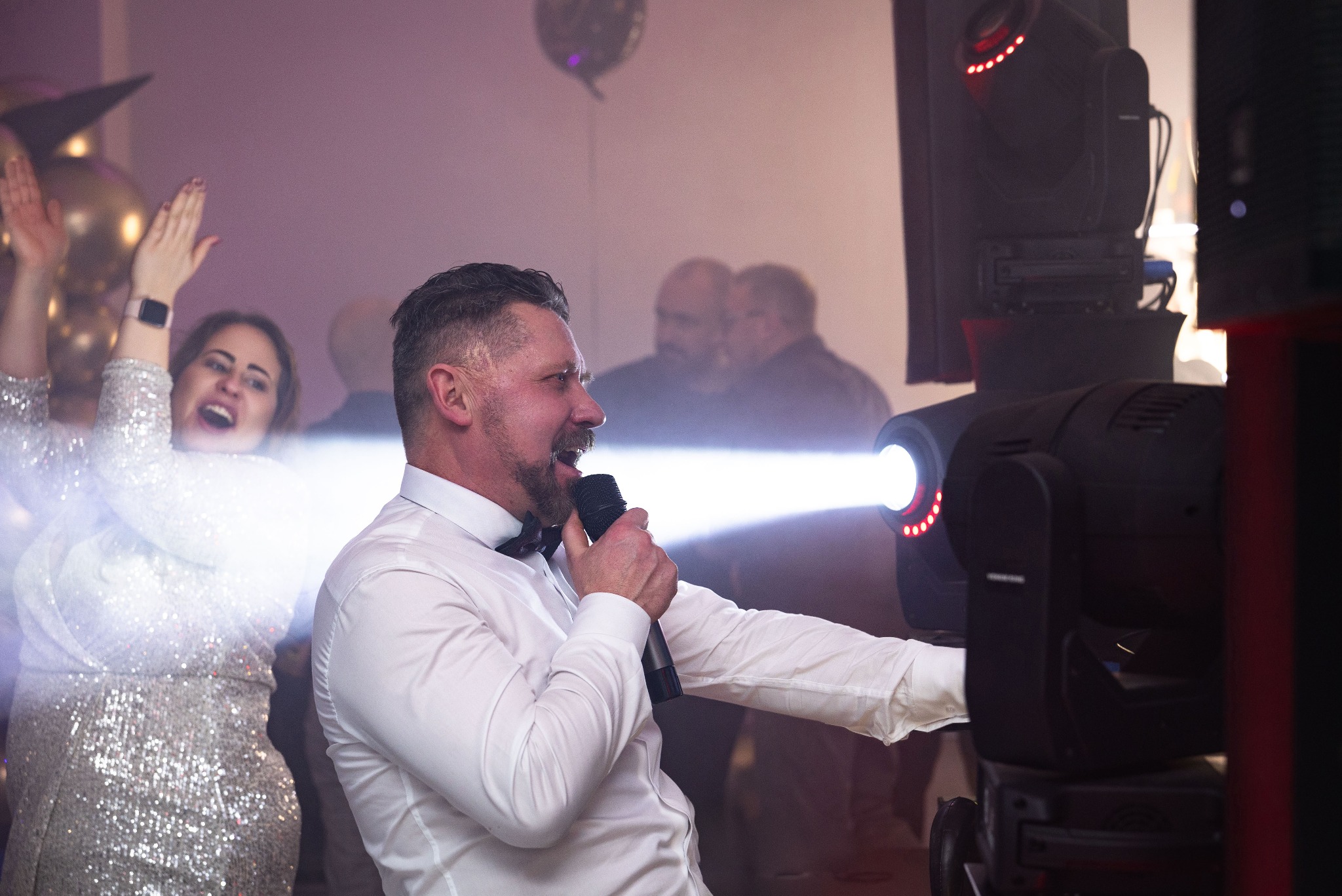Mężczyzna w białej koszuli i muszce śpiewa do mikrofonu na scenie podczas wydarzenia, w tle rozbawiona kobieta w cekinowej sukience i oświetlenie sceniczne.