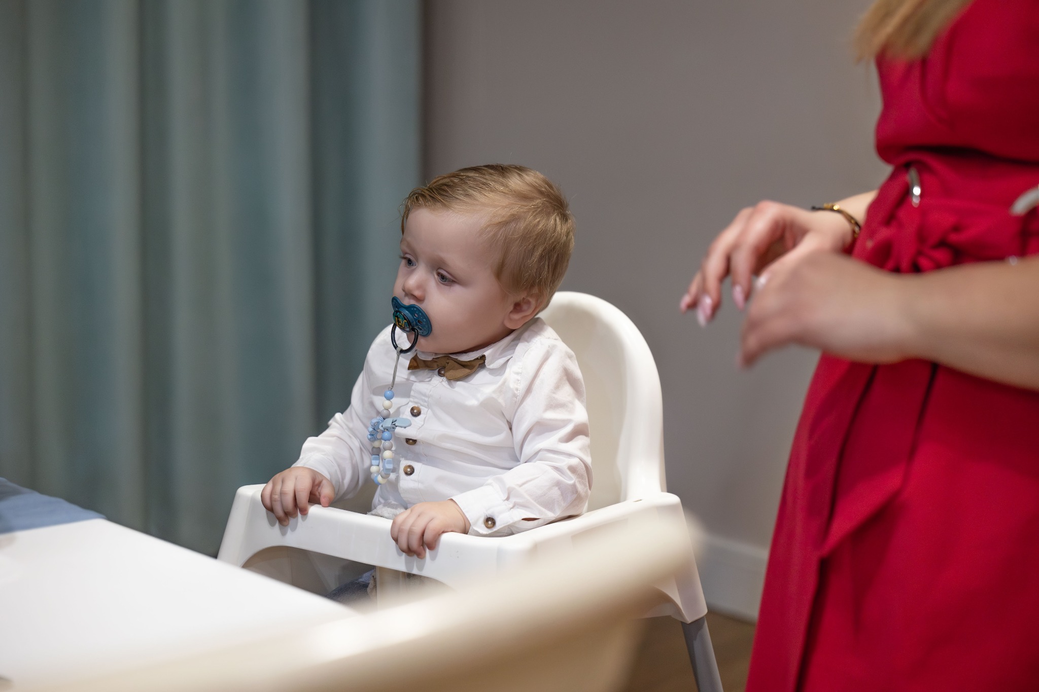 Chłopiec w białej koszuli i muszce siedzi w krzesełku do karmienia z smoczkiem w ustach, obok stoi kobieta w czerwonej sukience.