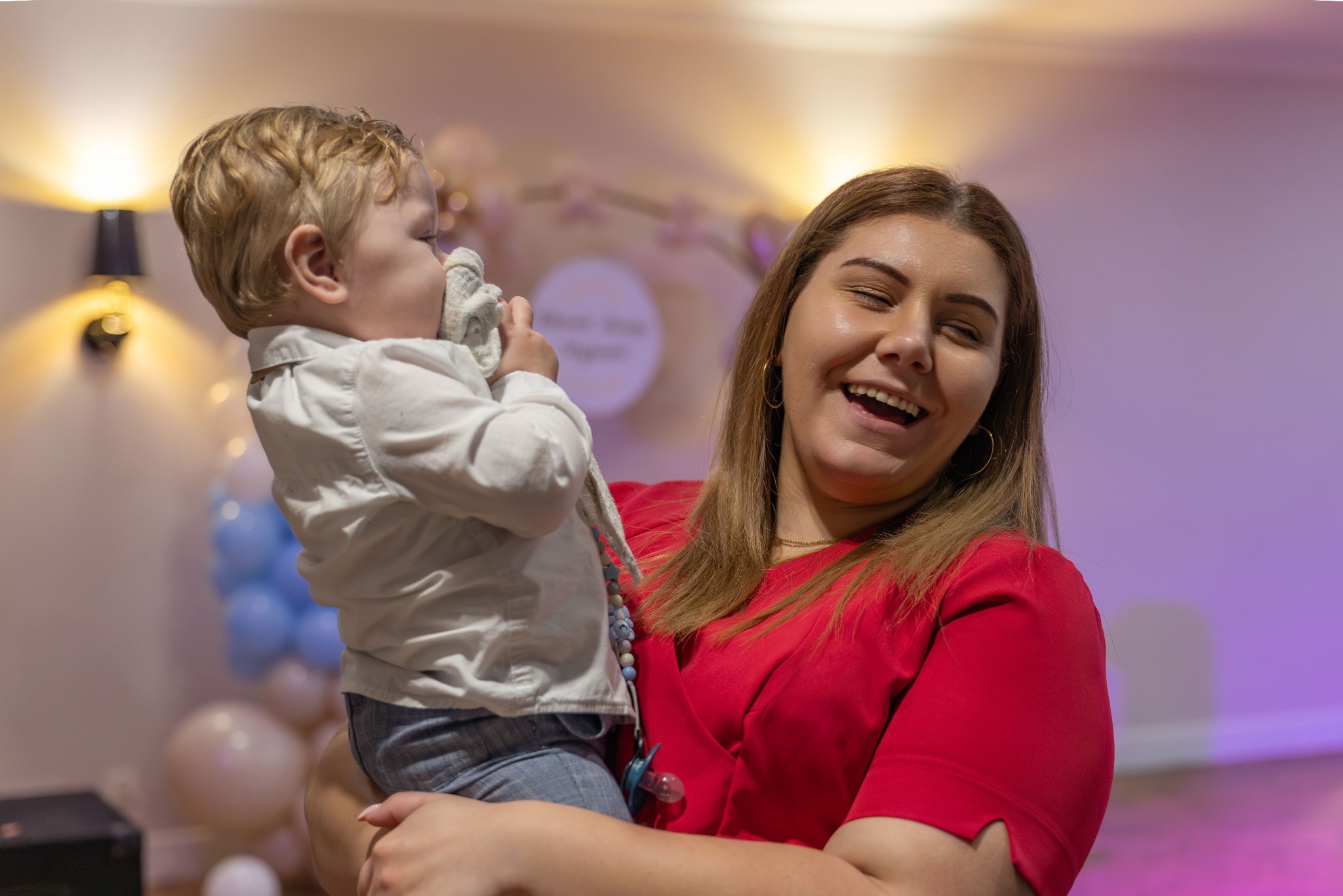 Uśmiechnięta kobieta w czerwonej sukience trzyma na rękach małego chłopca z blond włosami, który trzyma w buzi materiałową zabawkę, w tle dekoracje balonowe.