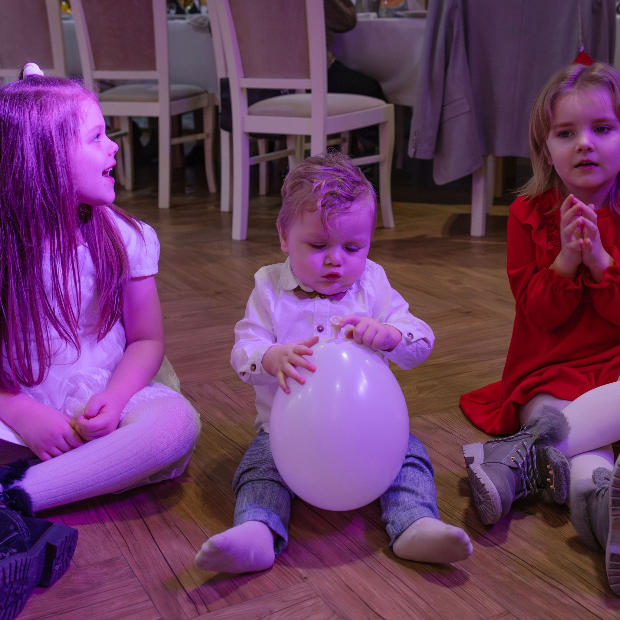 Troje dzieci siedzi na podłodze: dwie dziewczynki i chłopiec z białym balonem w rękach, oświetlenie sceniczne w odcieniach fioletu.