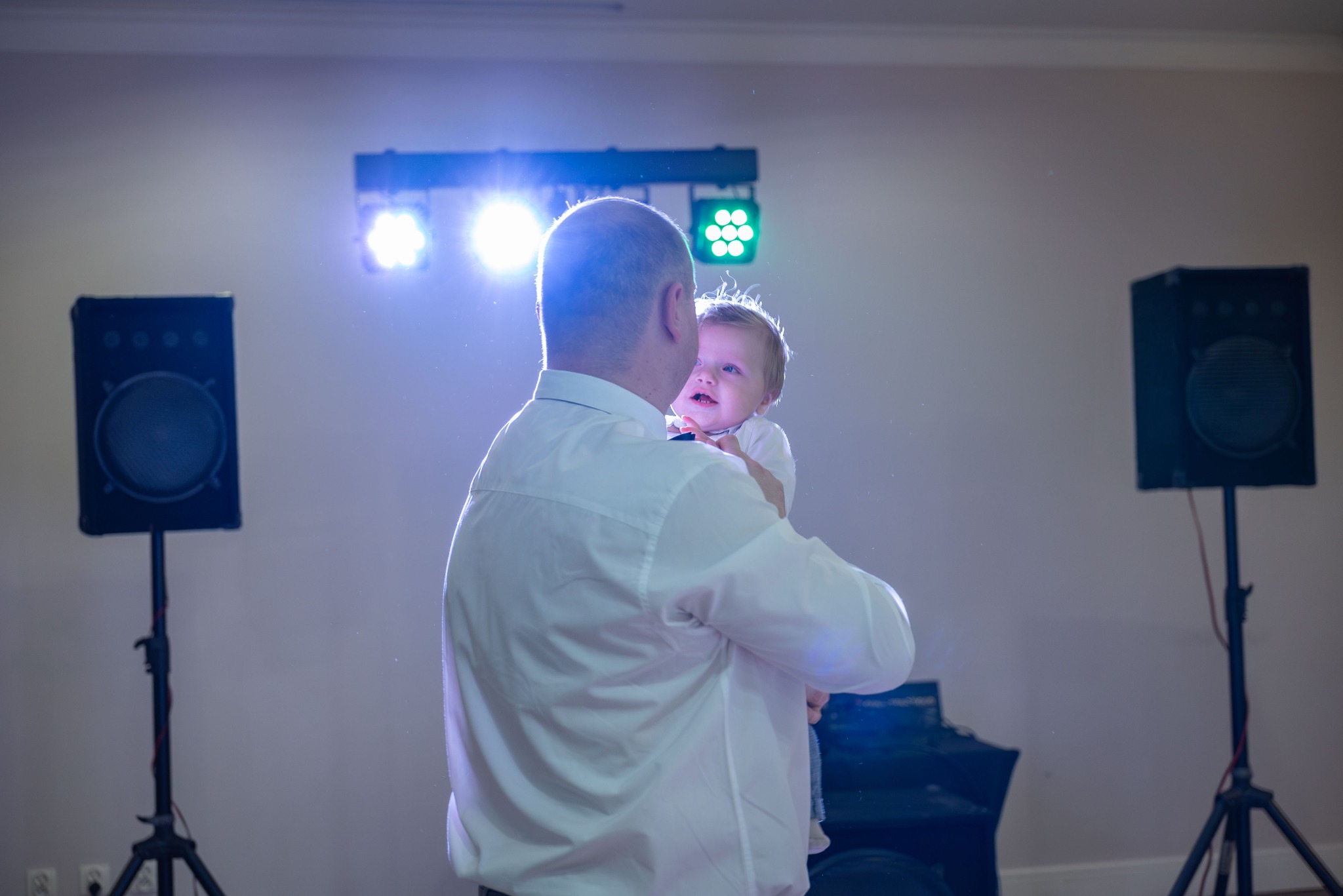 Ojciec trzymający małe dziecko na rękach na tle sceny z oświetleniem eventowym i głośnikami.