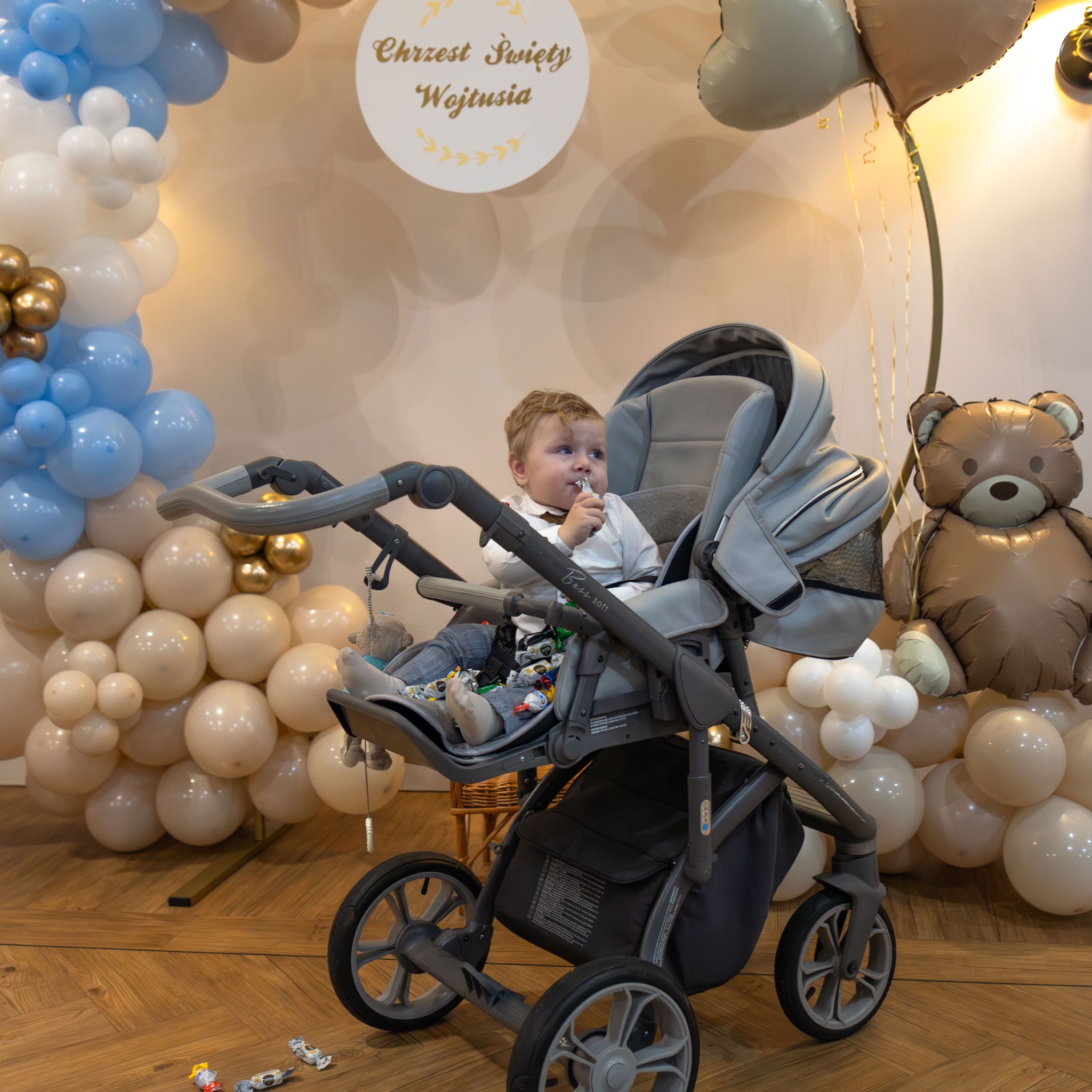 Chłopiec w szarej spacerówce na tle dekoracji balonowych z napisem 'Chrzest Święty Wojtusia', na podłodze leżą cukierki i zabawki.