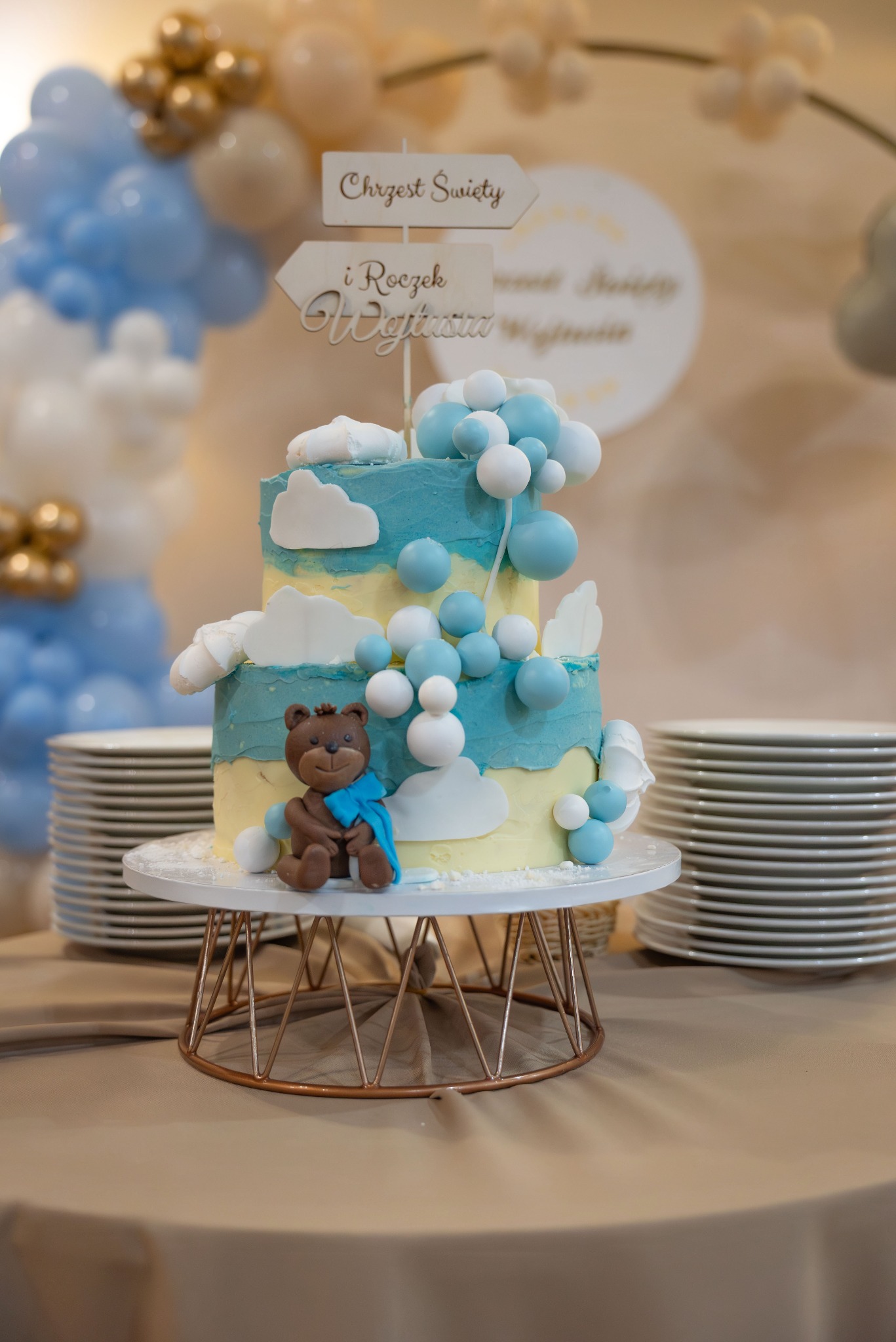 Dwupoziomowy tort zdobiony niebieskim kremem, białymi chmurkami z lukru i figurką misia z niebieską apaszką, dekoracja na roczek i chrzest święty, w tle balony i stosy talerzy.