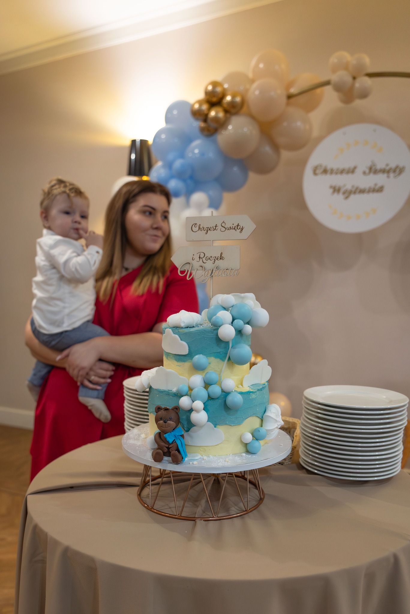 Dwupoziomowy tort w odcieniach błękitu i bieli, dekorowany chmurkami i kuleczkami, z małym czekoladowym misiem w niebieskiej apaszce, ustawiony na stole podczas uroczystości Chrztu Świętego, w tle...