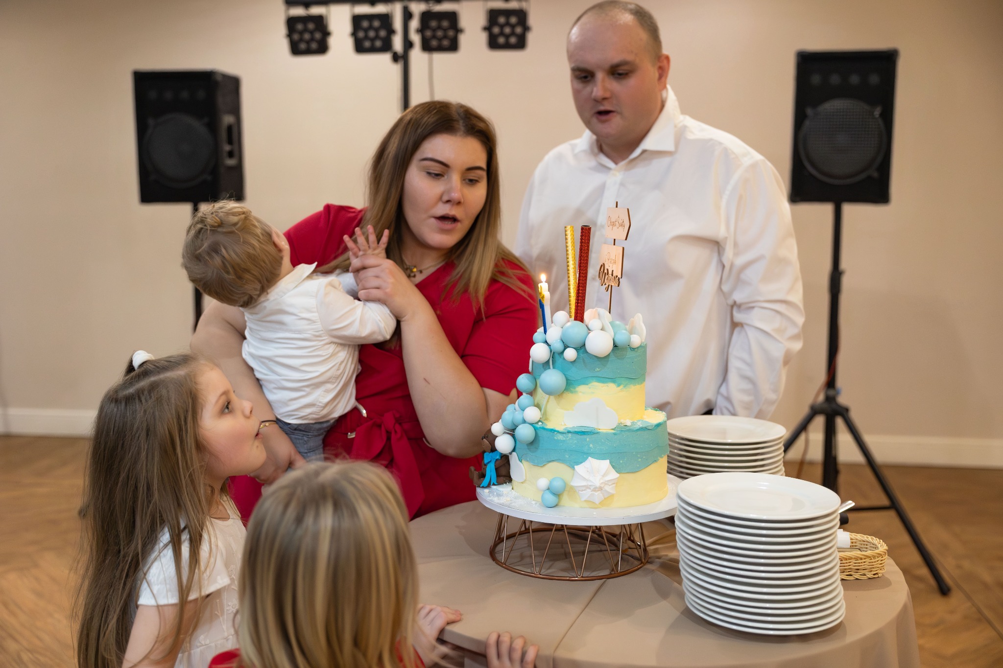 Rodzina z dziećmi świętuje urodziny z dwupoziomowym tortem w odcieniach błękitu i żółci, udekorowanym kulkami i świeczkami, w tle widoczny sprzęt nagłośnieniowy.