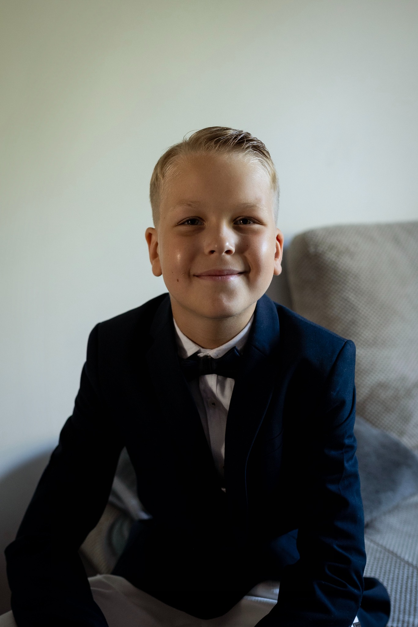 Uśmiechnięty chłopiec w granatowej marynarce i muszce, siedzący na szarej sofie na tle jasnej ściany.