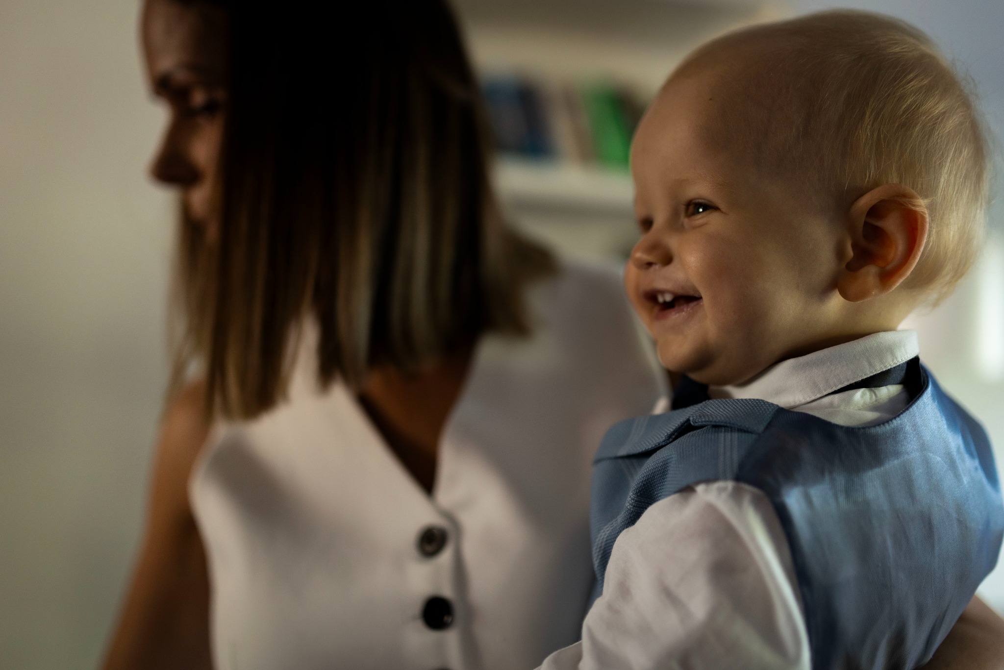 Uśmiechnięty blond chłopiec w niebieskiej kamizelce, trzymany na rękach przez kobietę w białej bluzce, z zielonymi książkami w tle.