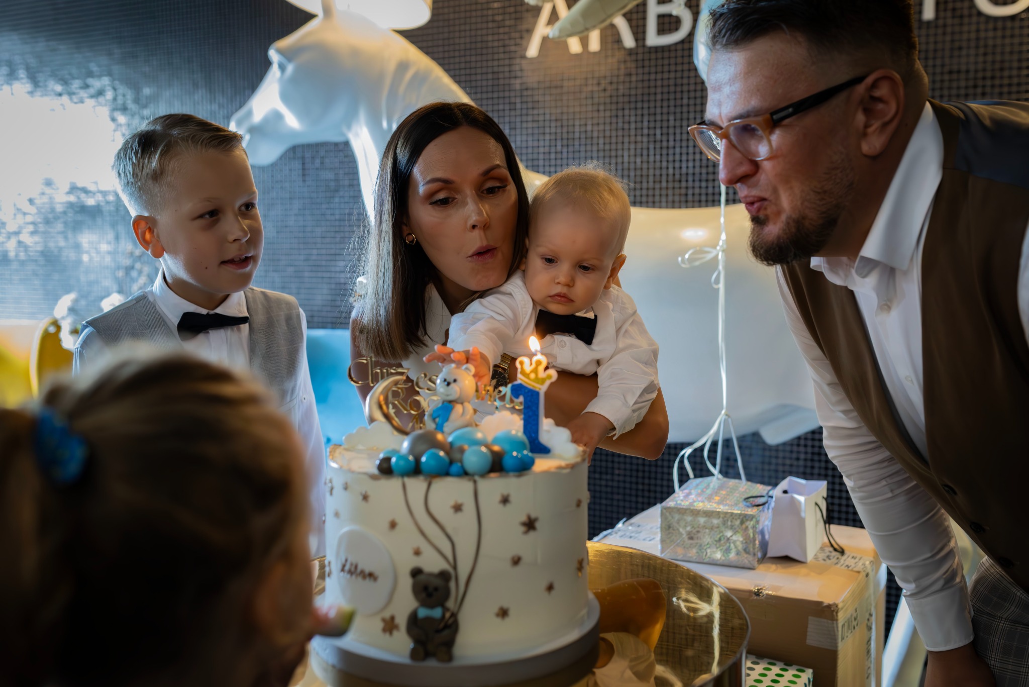 Rodzina z tortem urodzinowym ozdobionym cyfrą jeden i figurką misia, mama trzyma dziecko, tata zamierza zdmuchnąć świeczkę, w tle dekoracja w kształcie białego konia.