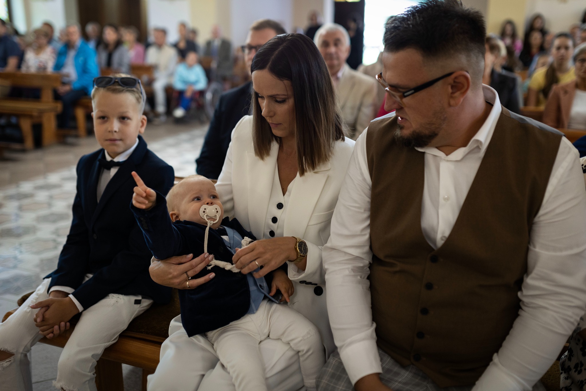 Rodzina w kościele podczas uroczystości: matka w białym garniturze trzyma niemowlę z smoczkiem, obok siedzi starszy syn w eleganckim ubraniu i ojciec w kamizelce, w tle zgromadzeni ludzie na ławkach.