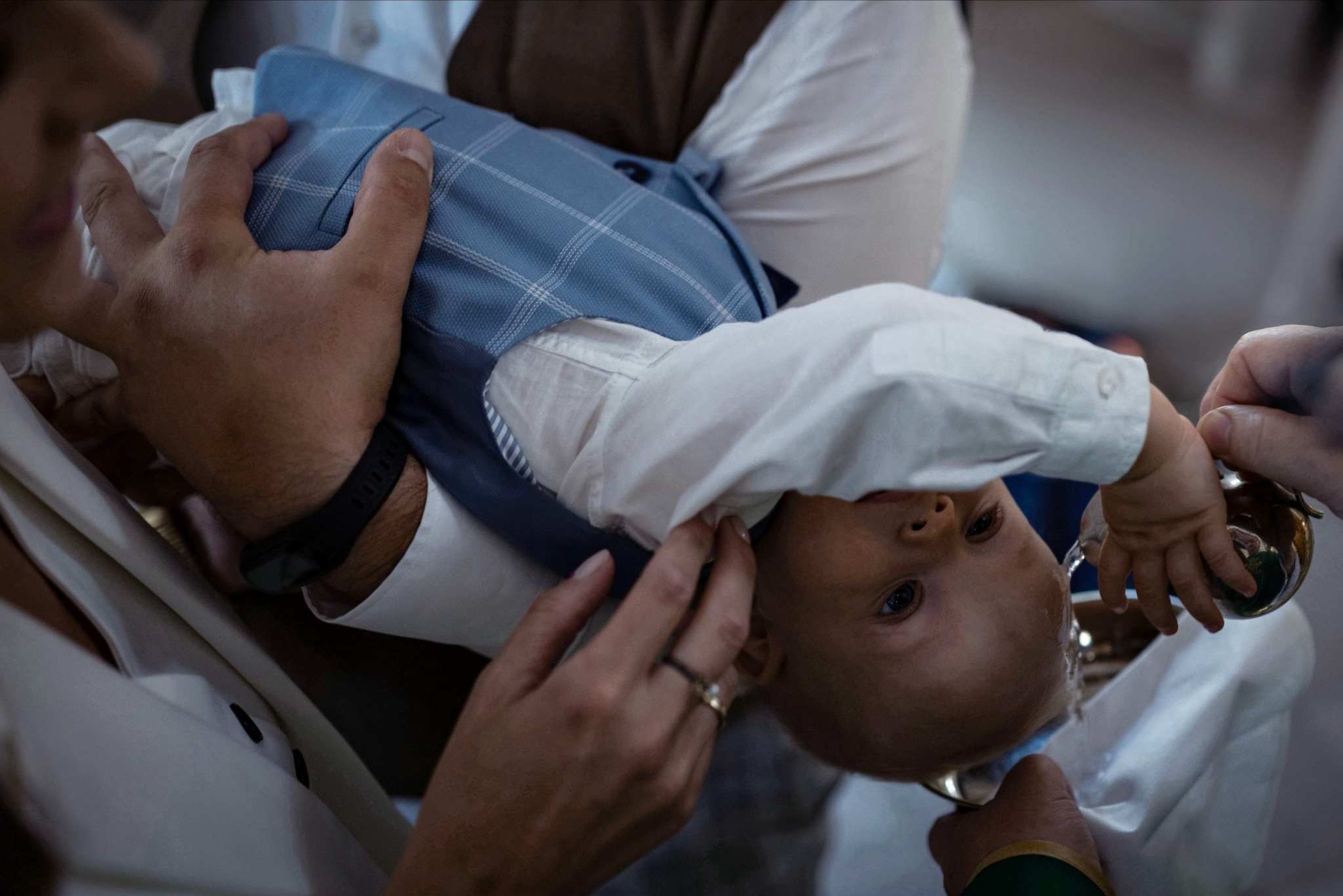 Niemowlę w niebieskiej kamizelce, trzymane przez dorosłych, podczas ceremonii z użyciem srebrnego naczynia i wody.